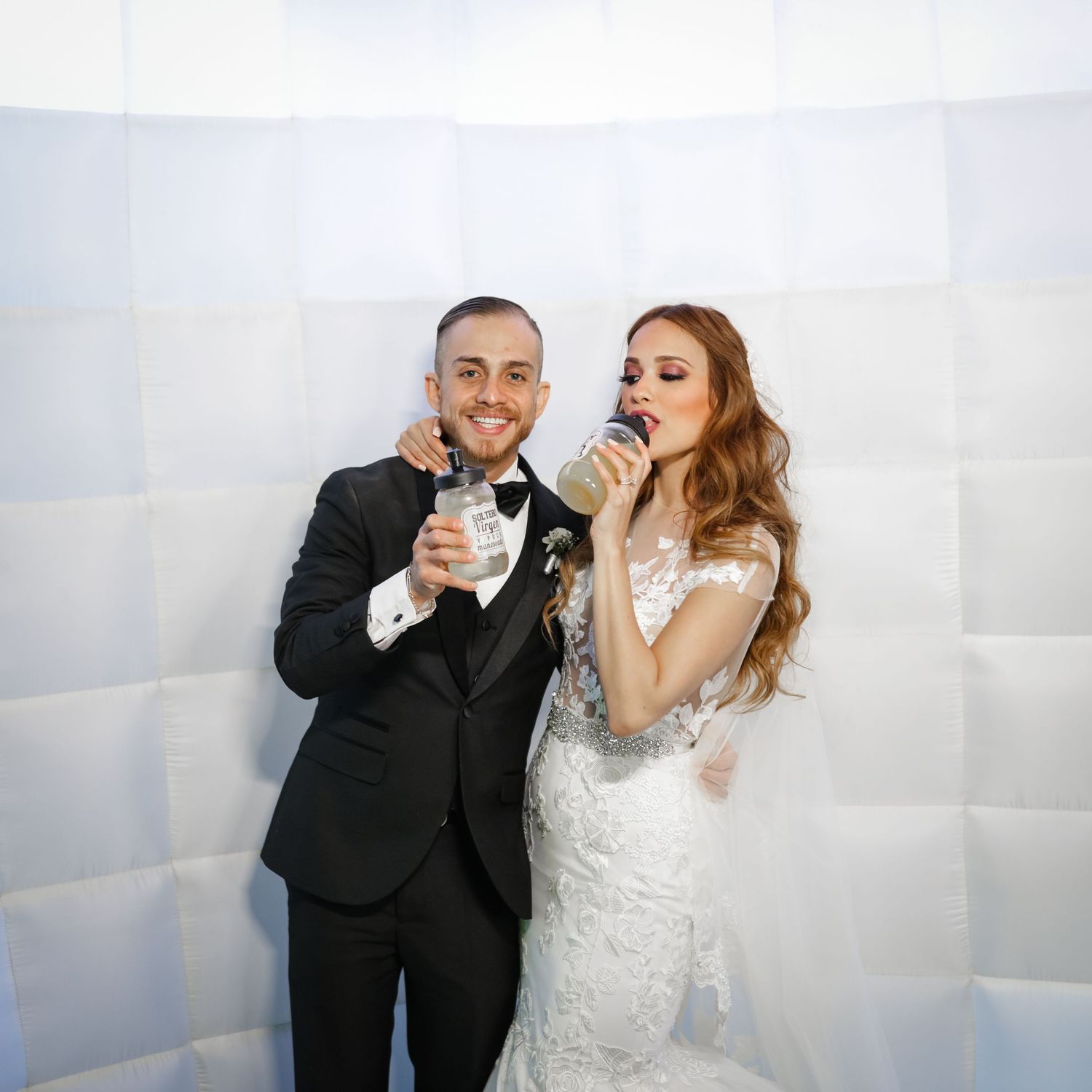 Smiling bride and groom pose in a photo booth, pretending to drink from silver cans together.