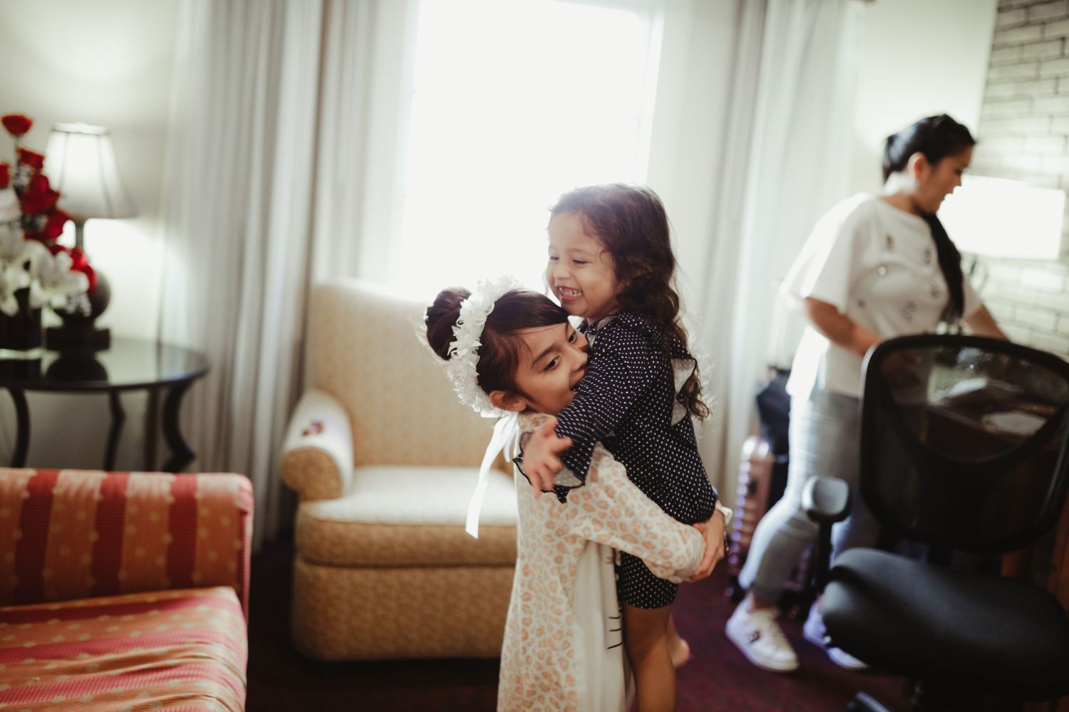 Two young girls share a joyful hug in a hotel room during wedding day preparations.