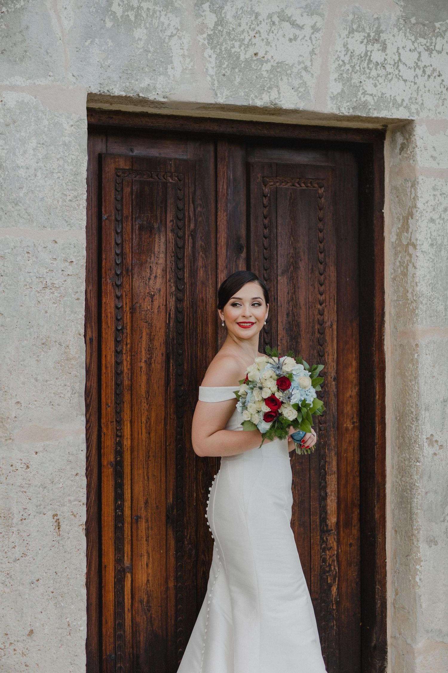 Elegant bride in off-shoulder white gown holding floral bouquet standing before ornate wooden doors.