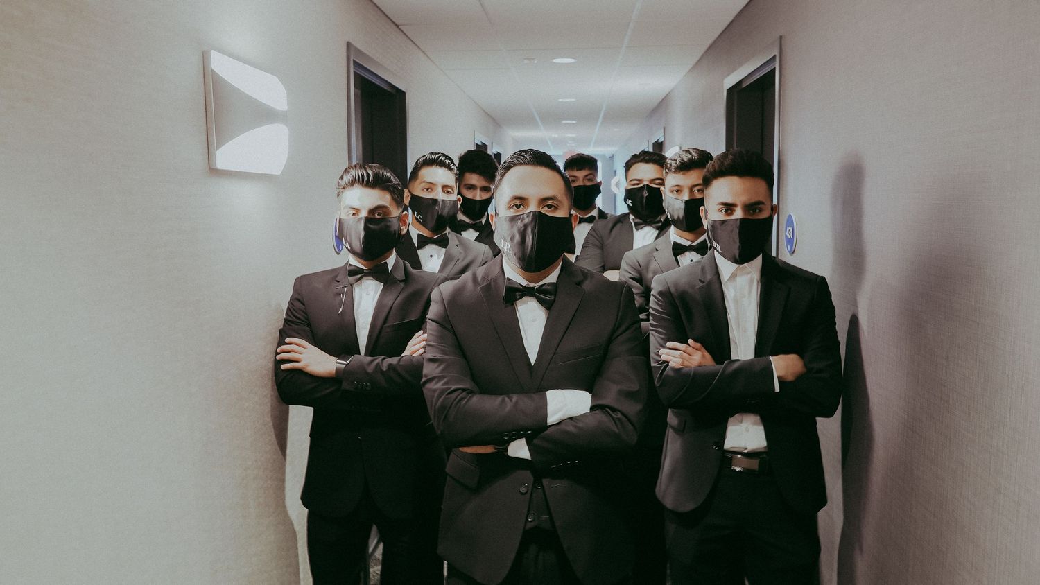 Group of men in formal black tuxedos and bow ties wearing black face masks standing confidently in a hallway.