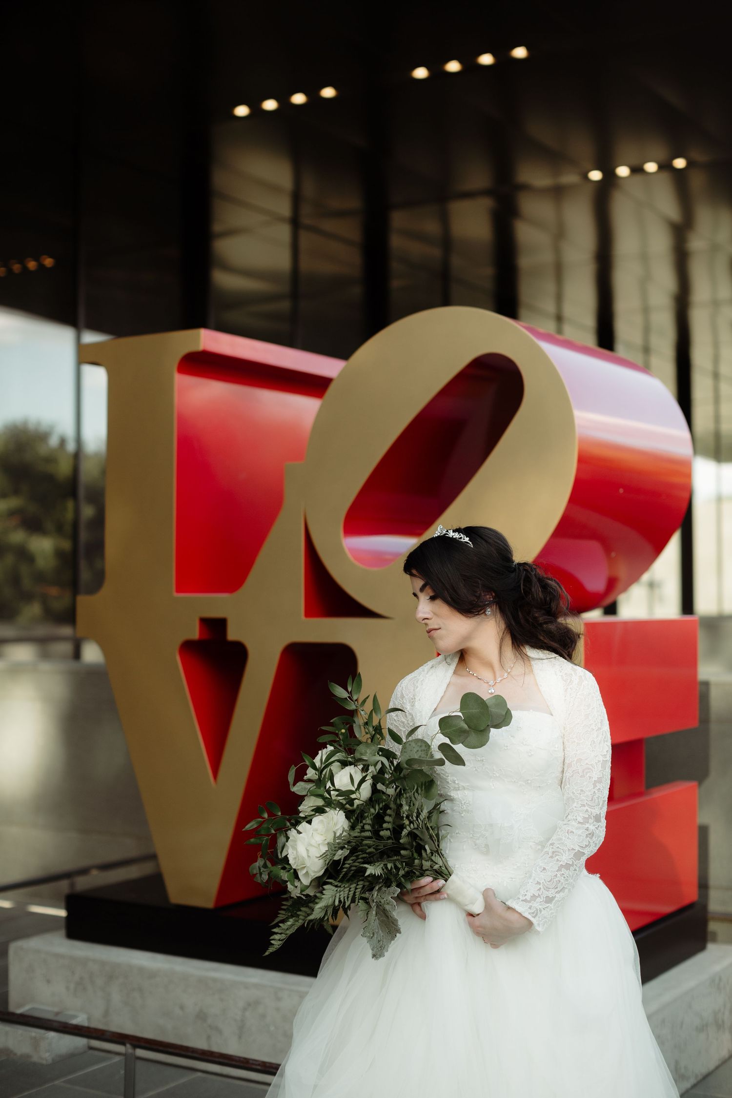 Bride in white lace gown holding bouquet posing before large red LOVE sculpture.