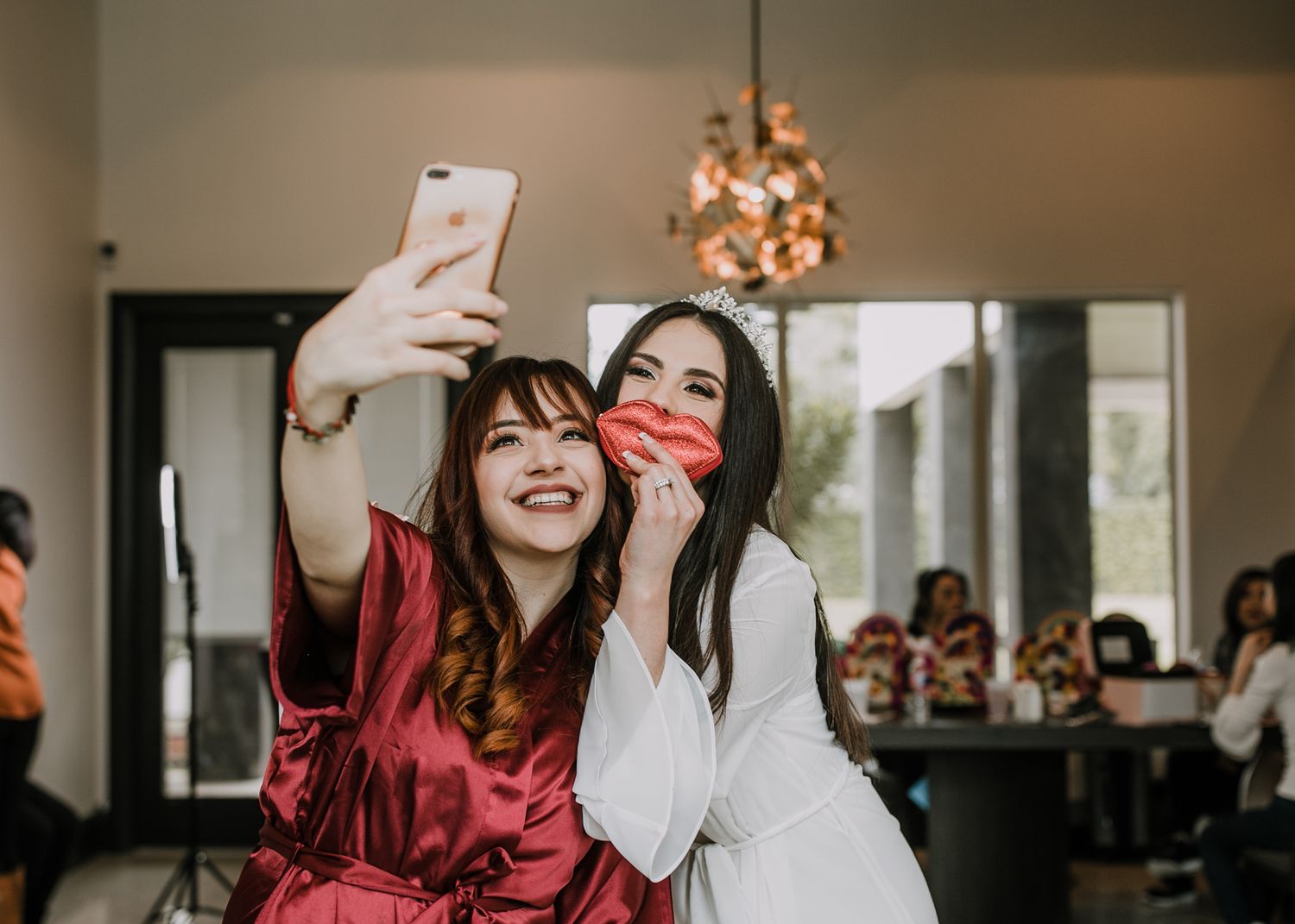 Two women taking selfie during bridal party preparations, one in red robe.