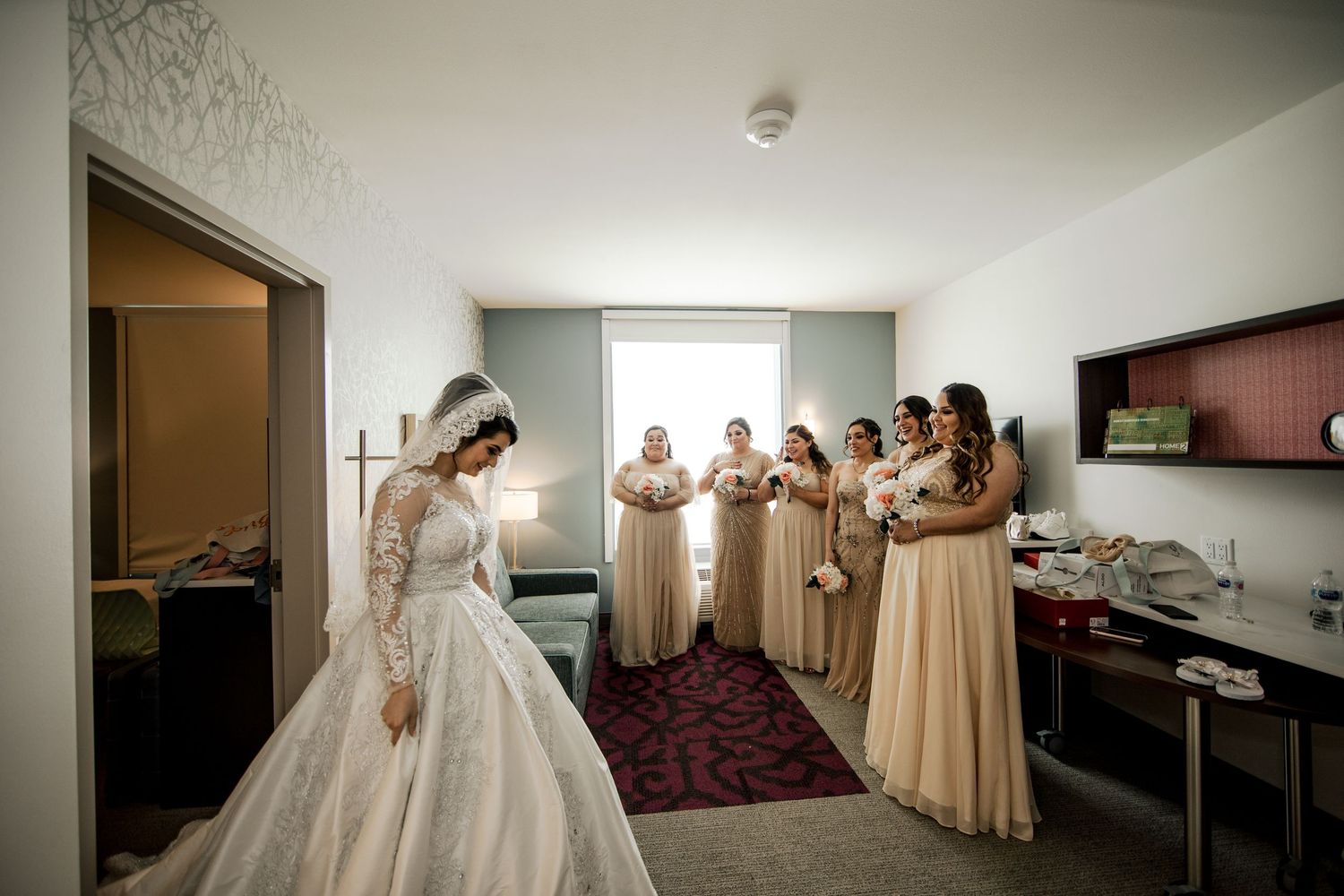 Bride in elegant lace gown reveals dress to bridesmaids in champagne dresses in hotel room.