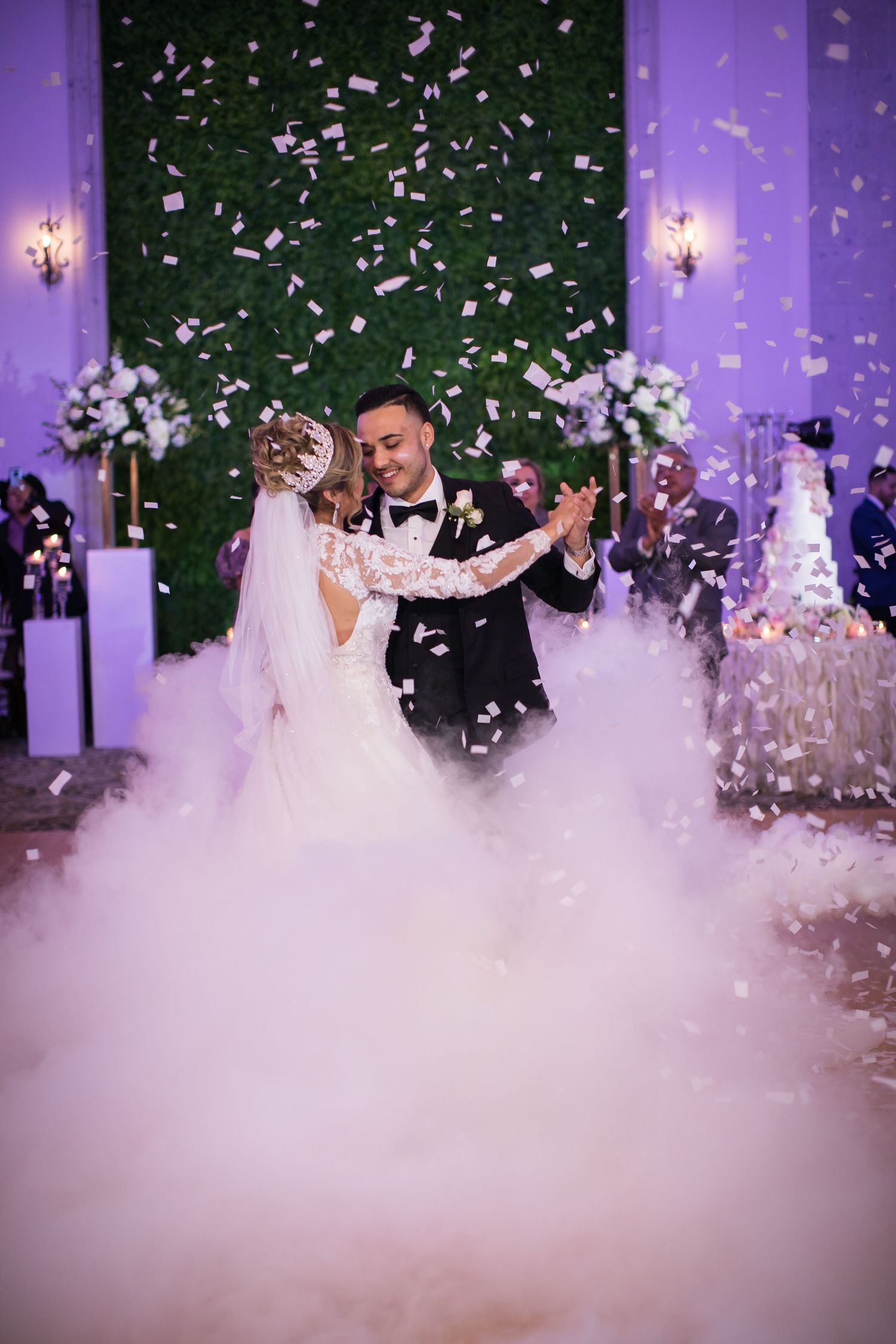 Bride and groom share first dance surrounded by dramatic fog and falling confetti at wedding reception.