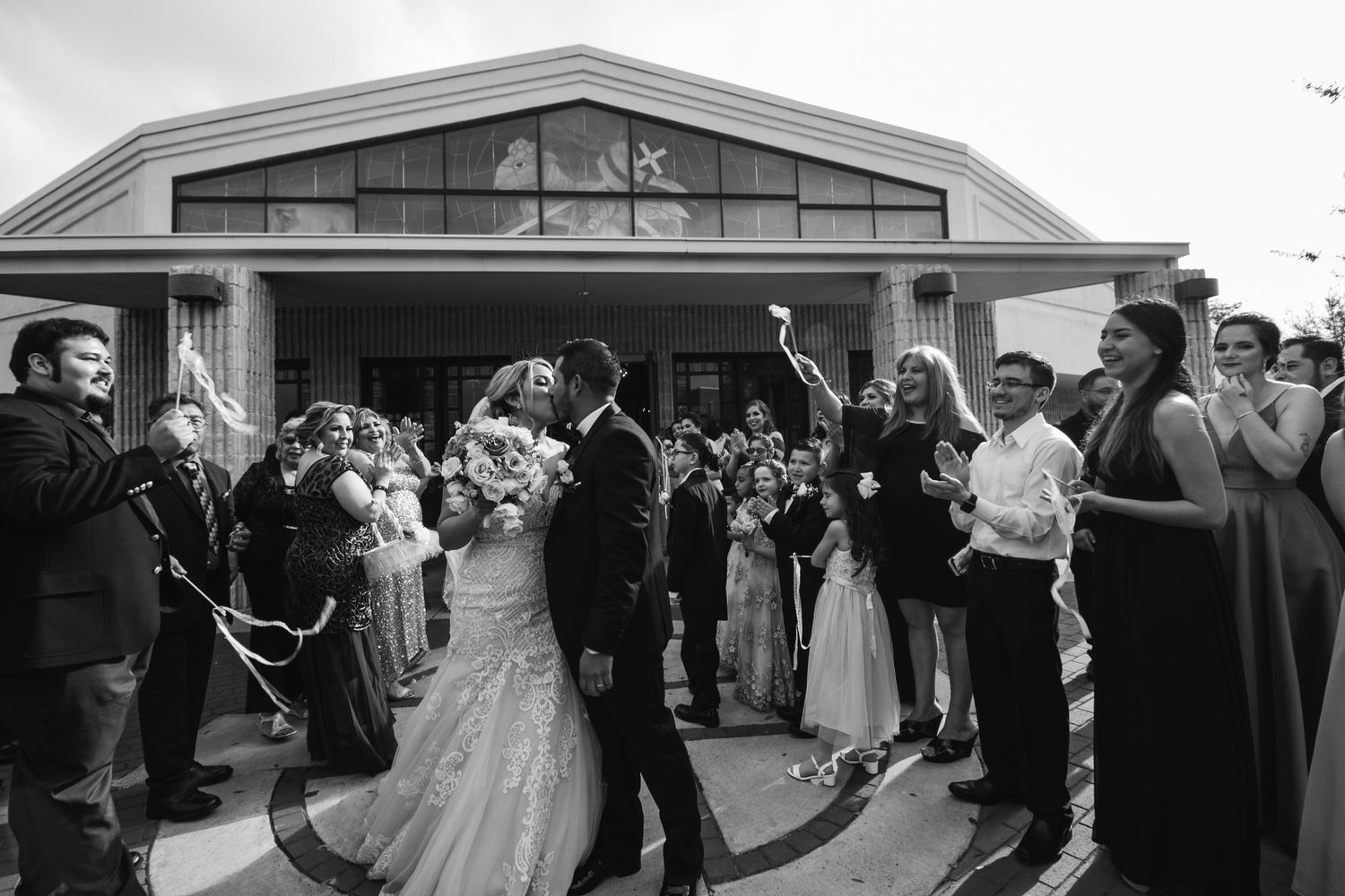 Black and white photo of newlyweds kissing as guests celebrate their church exit with streamers.