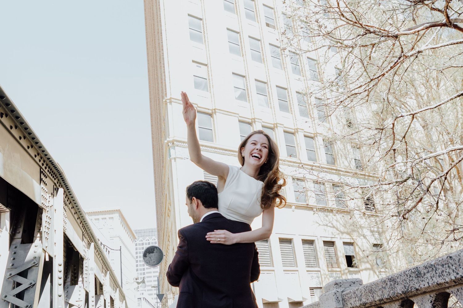 A joyful couple celebrates outdoors, man lifting laughing woman near urban building with blooming trees.