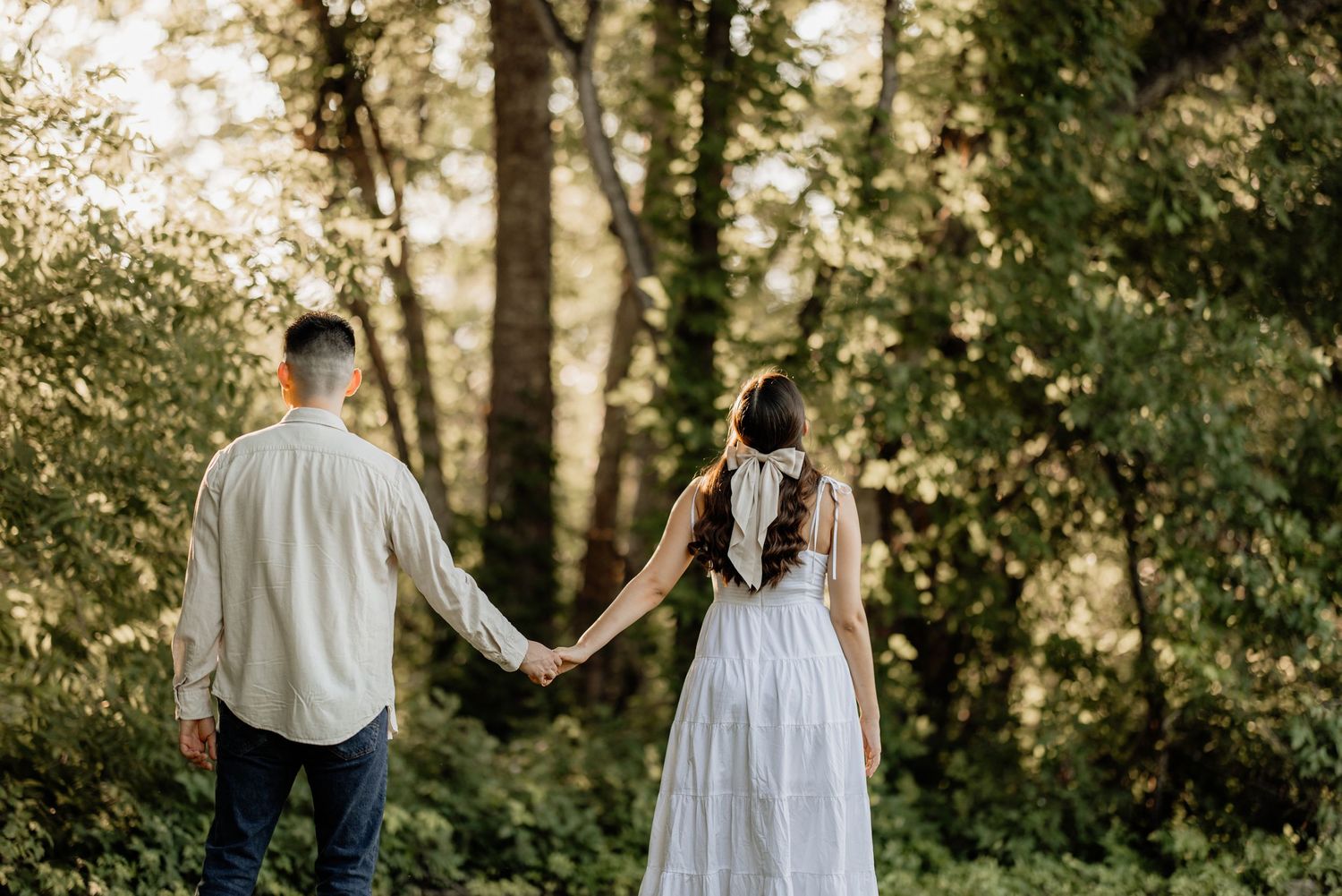 Couple holding hands walking toward sunlit forest in romantic outdoor engagement photo session.