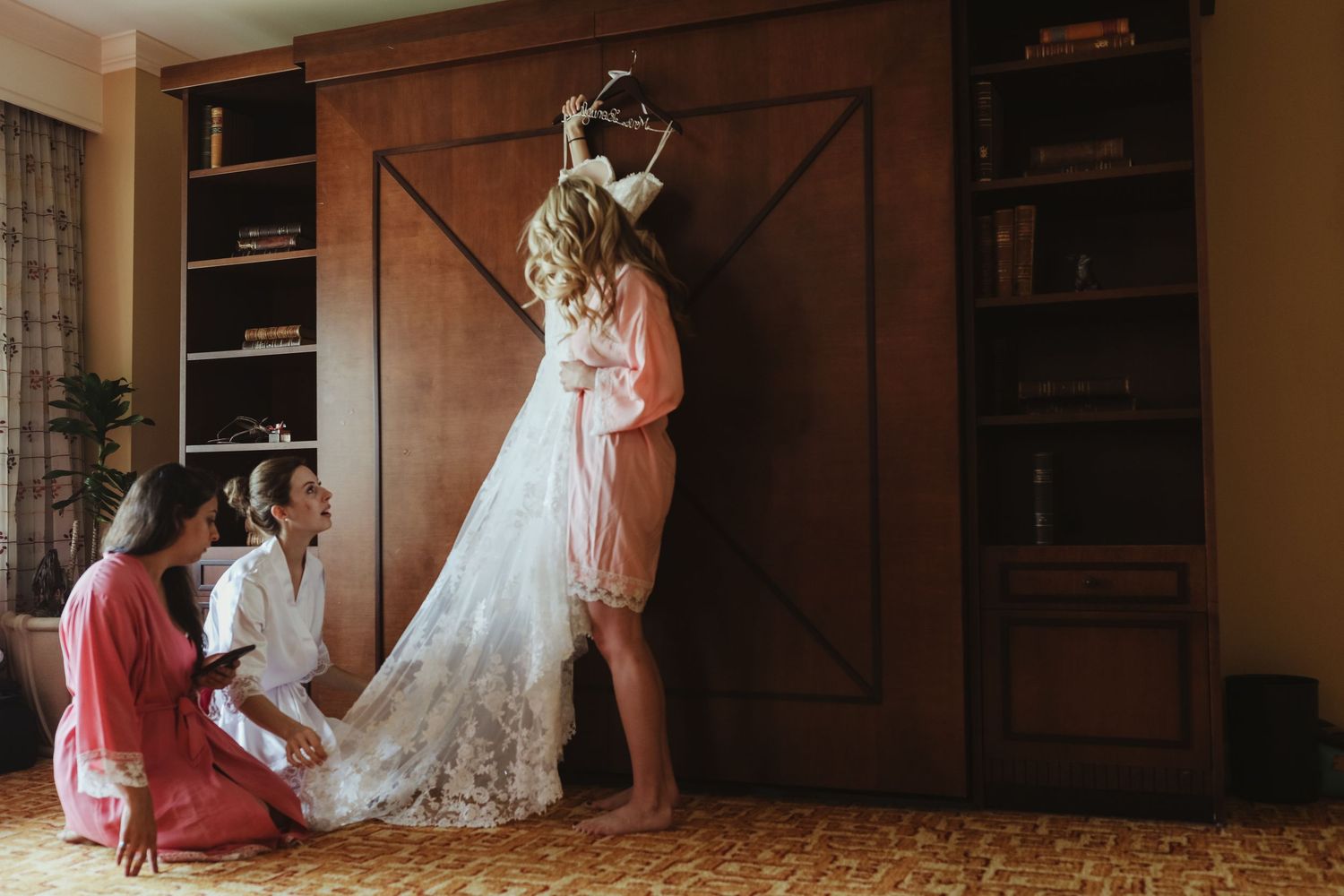 Bridesmaids help bride into elegant lace wedding gown in warmly decorated bridal suite before ceremony.