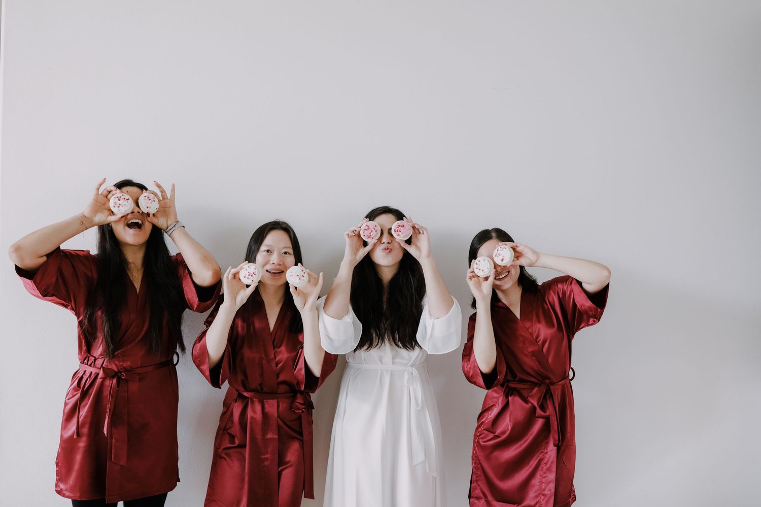 Four bridesmaids in red satin robes hold macarons over their eyes while posing with bride in white robe.