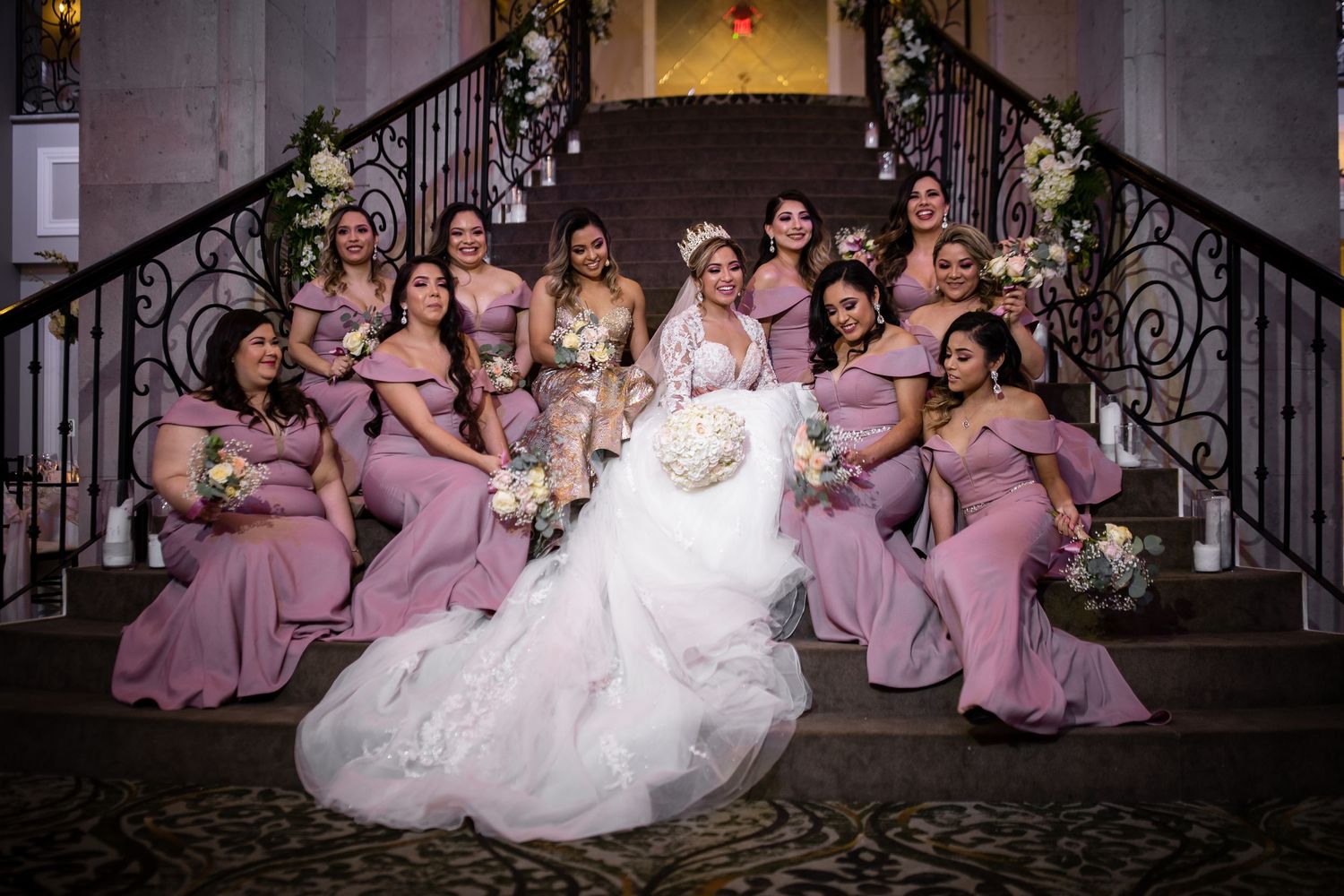 Bride surrounded by bridesmaids in dusty rose gowns posing on grand staircase with floral decor.