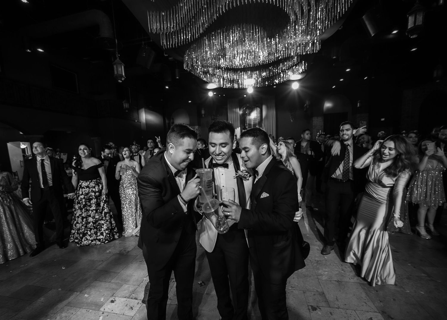 Three men in suits celebrate together on a dance floor at a wedding reception in black and white.