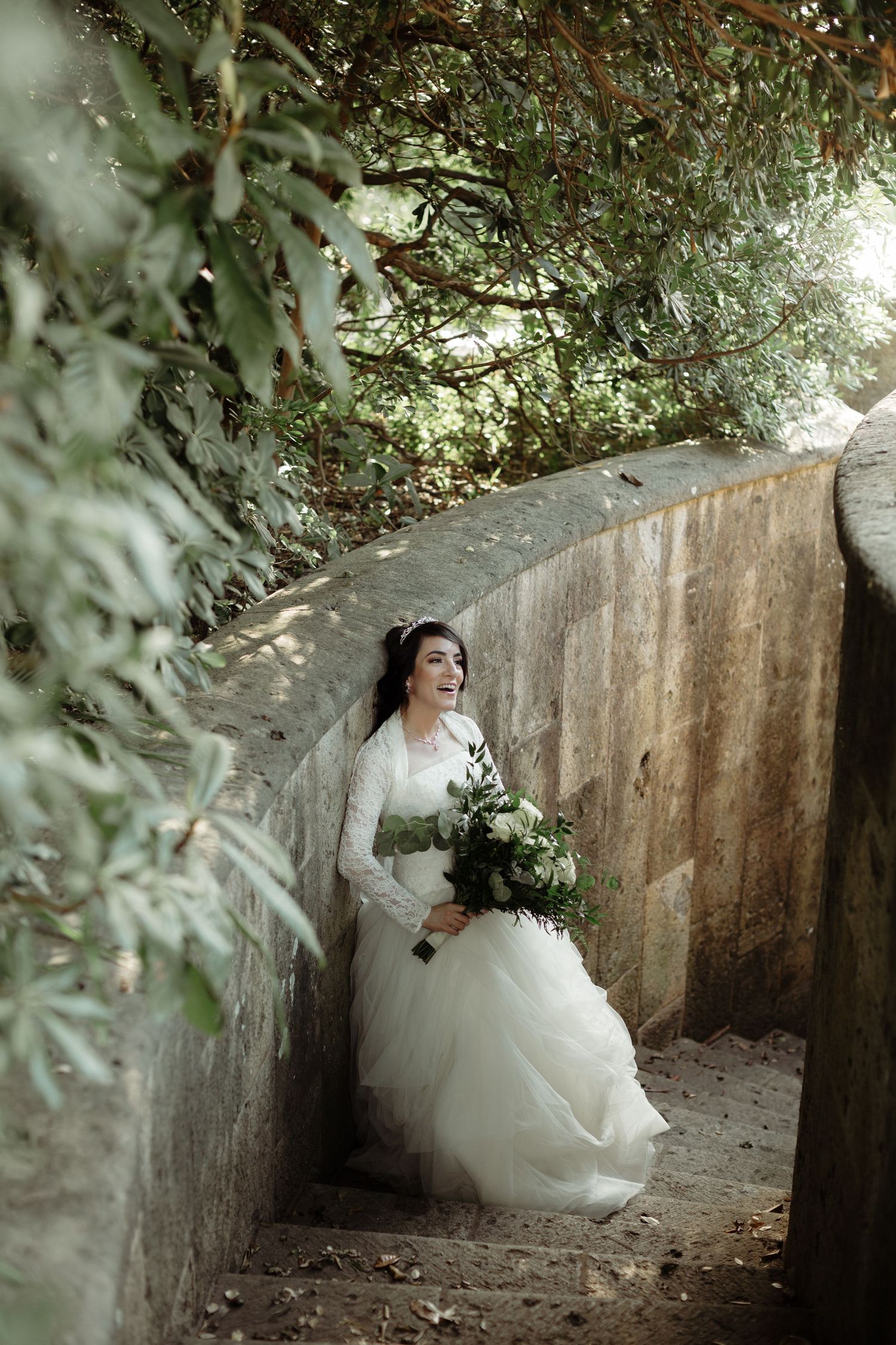Bride in white lace gown holding greenery bouquet smiling in a stone stairway surrounded by lush foliage.