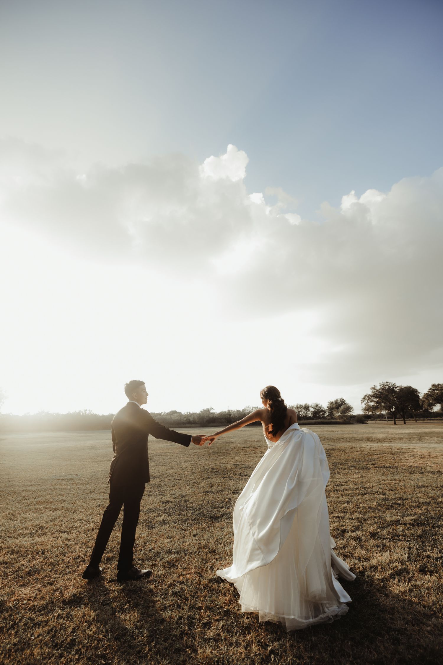 Bride and groom holding hands in a sunlit field, bride in flowing white gown, golden hour wedding photo.