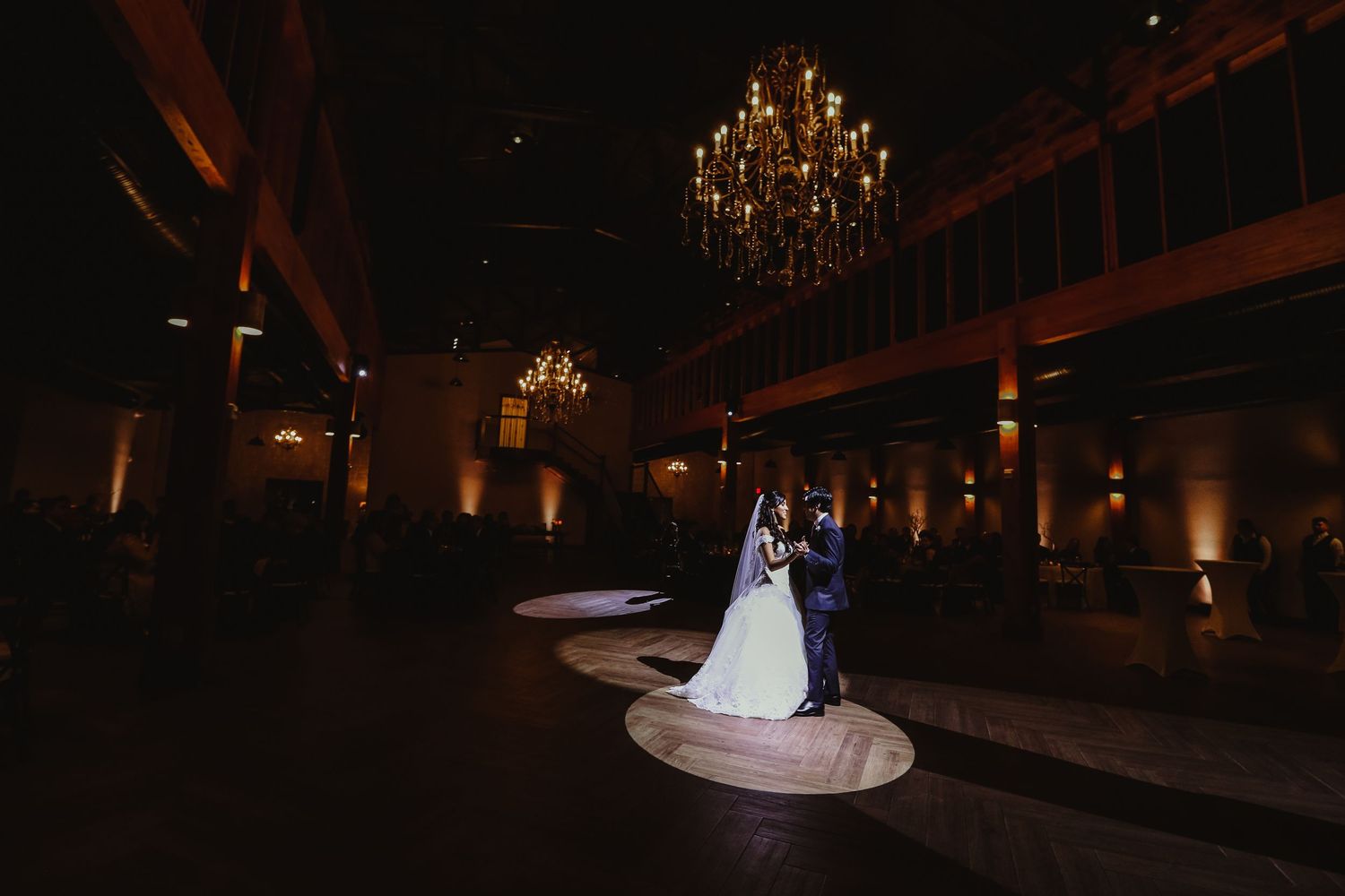 Bride and groom share their first dance under a grand chandelier in a dramatically lit wedding reception venue.