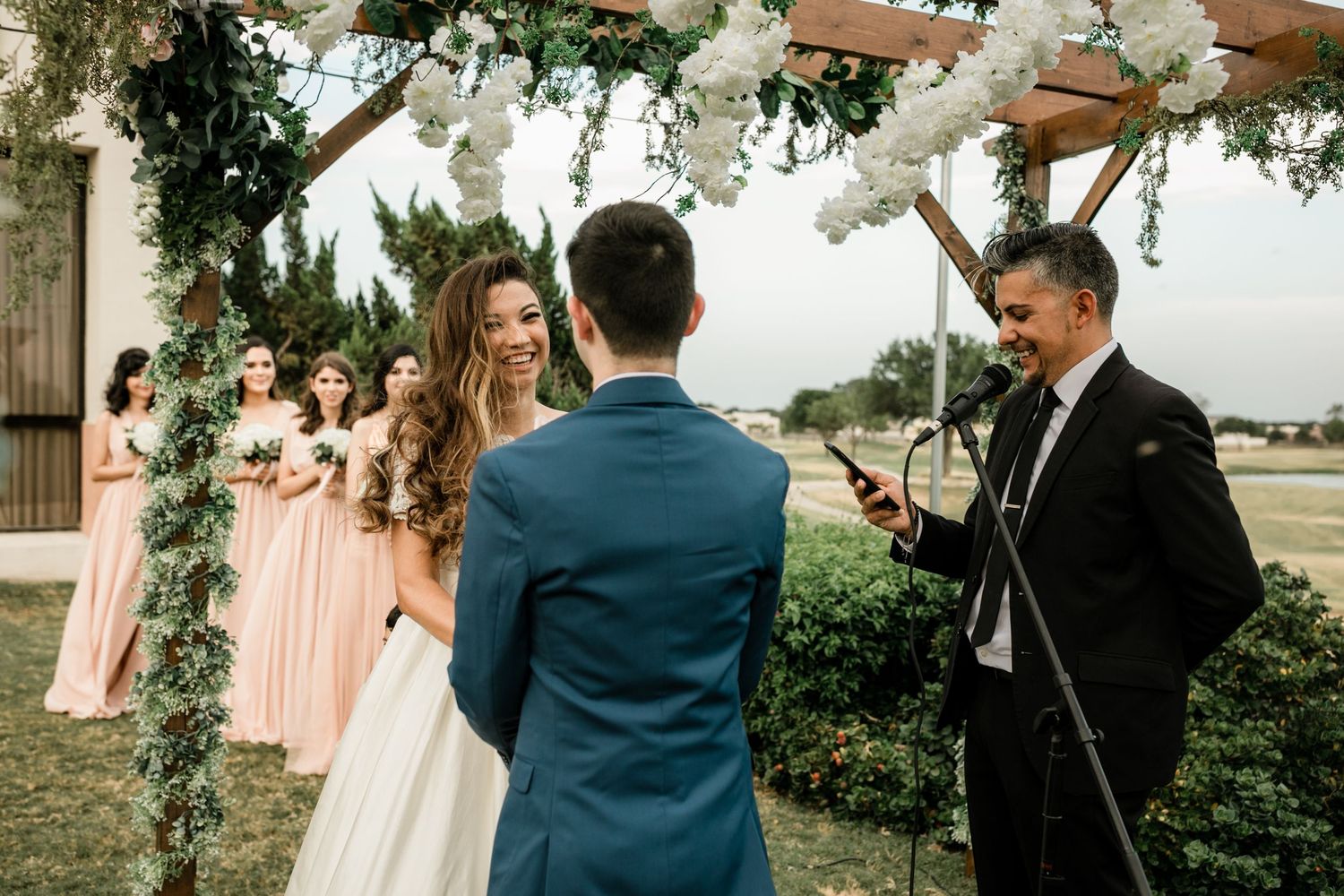 Bride and groom exchange vows under floral arch with bridesmaids in blush dresses nearby.