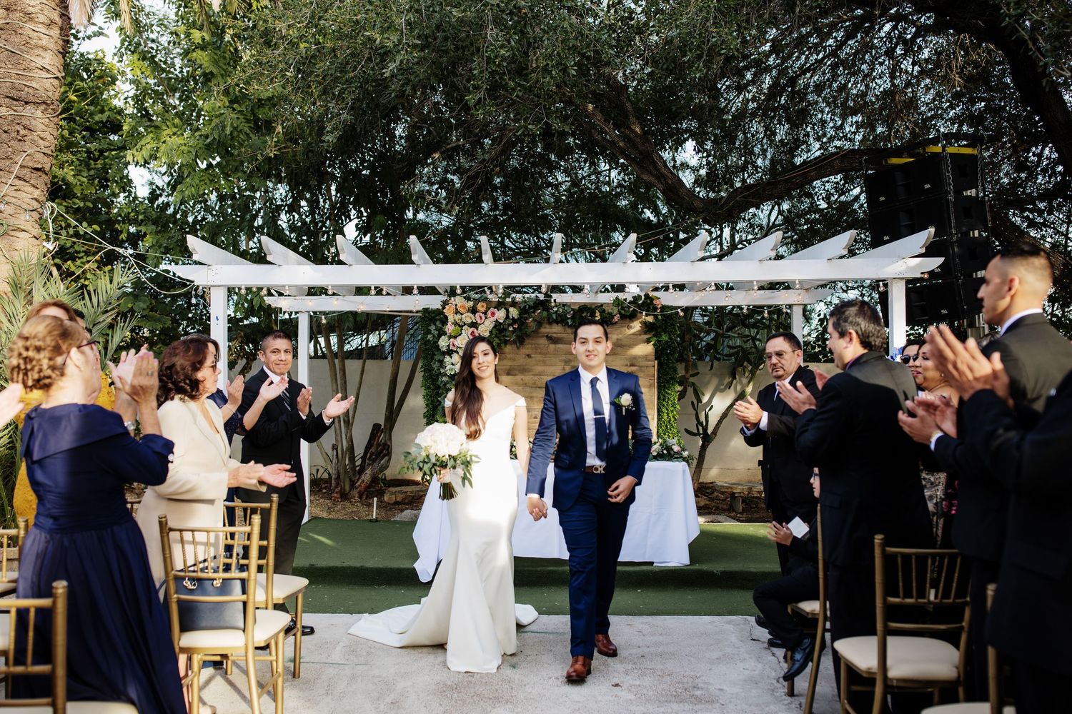 Newlyweds walk down outdoor aisle as guests applaud beneath white floral wedding canopy.