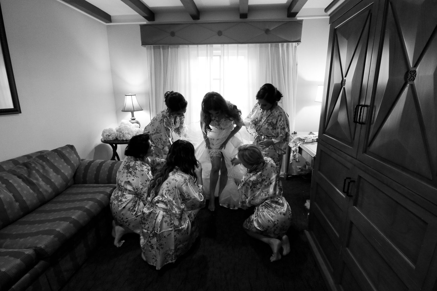 Bridesmaids in floral robes gather around bride during wedding preparations in a hotel room.