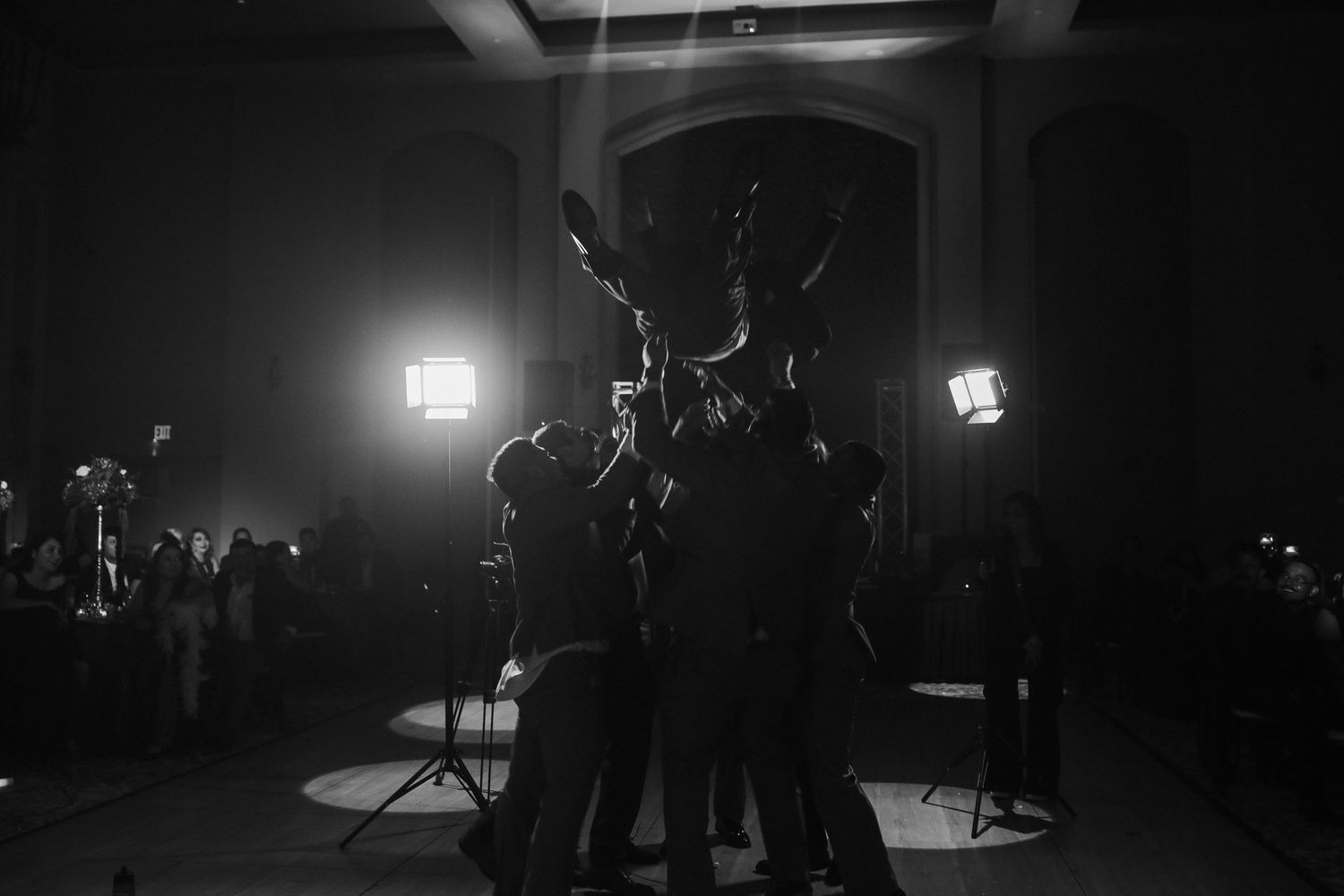 Black and white photo of wedding guests celebrating on a dark dance floor with bright stage lights.