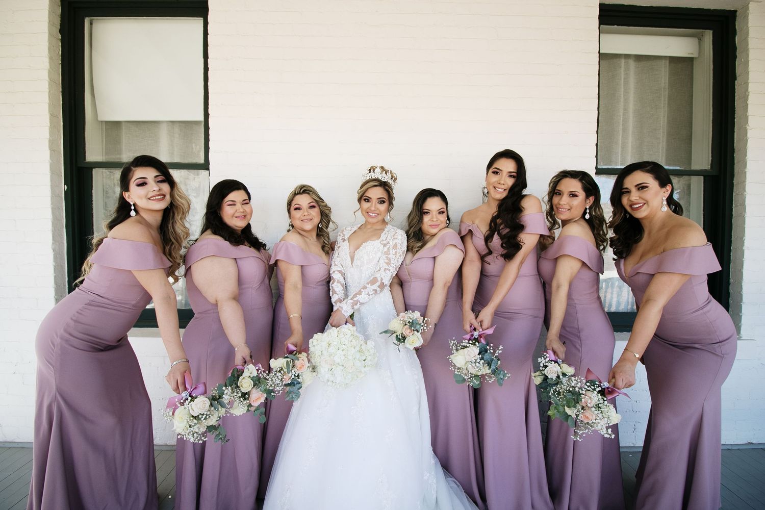 Bride wearing crown with seven bridesmaids in dusty purple off-shoulder gowns holding bouquets.