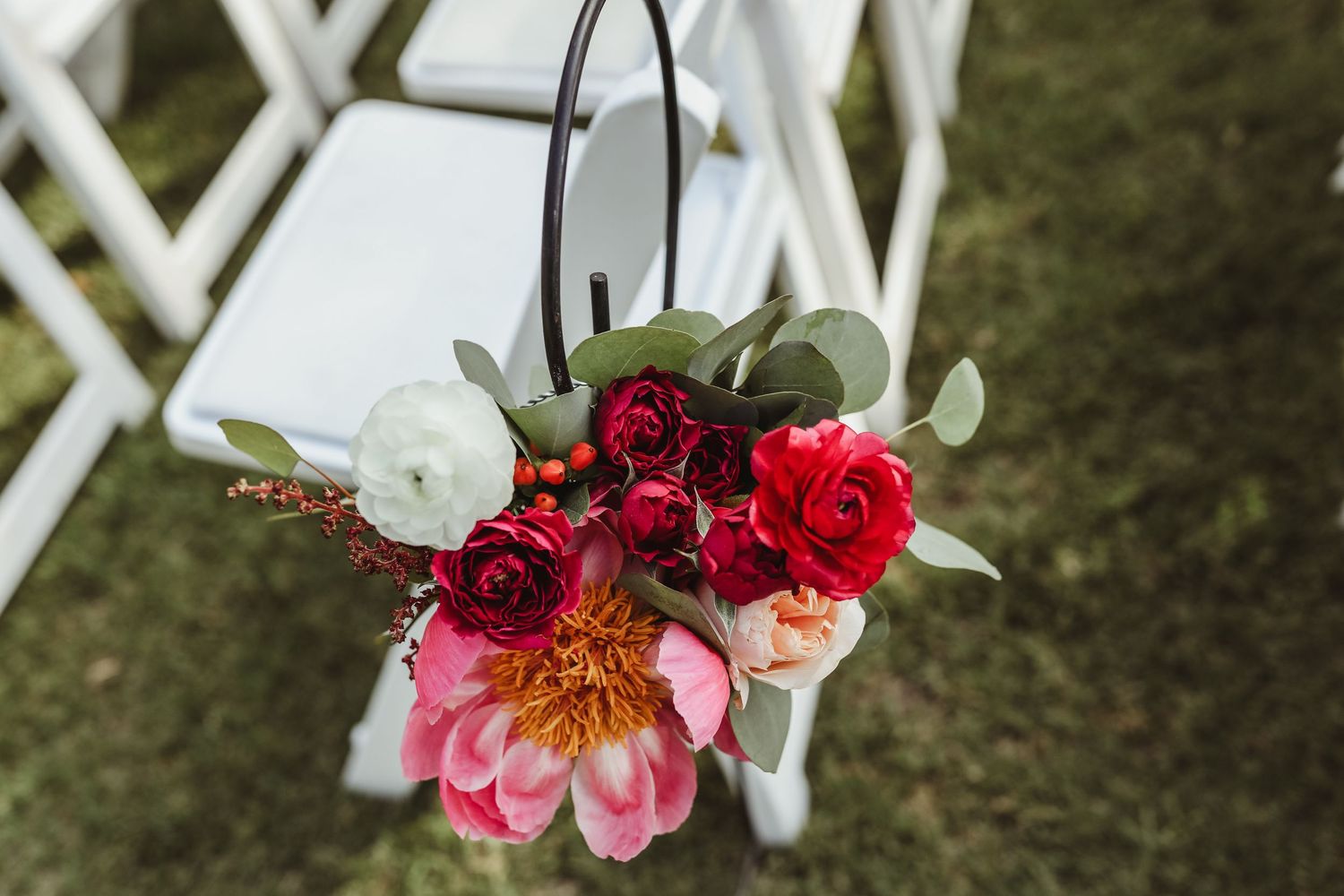 Vibrant red, pink, and white floral arrangement hanging on a white wedding ceremony chair outdoors.