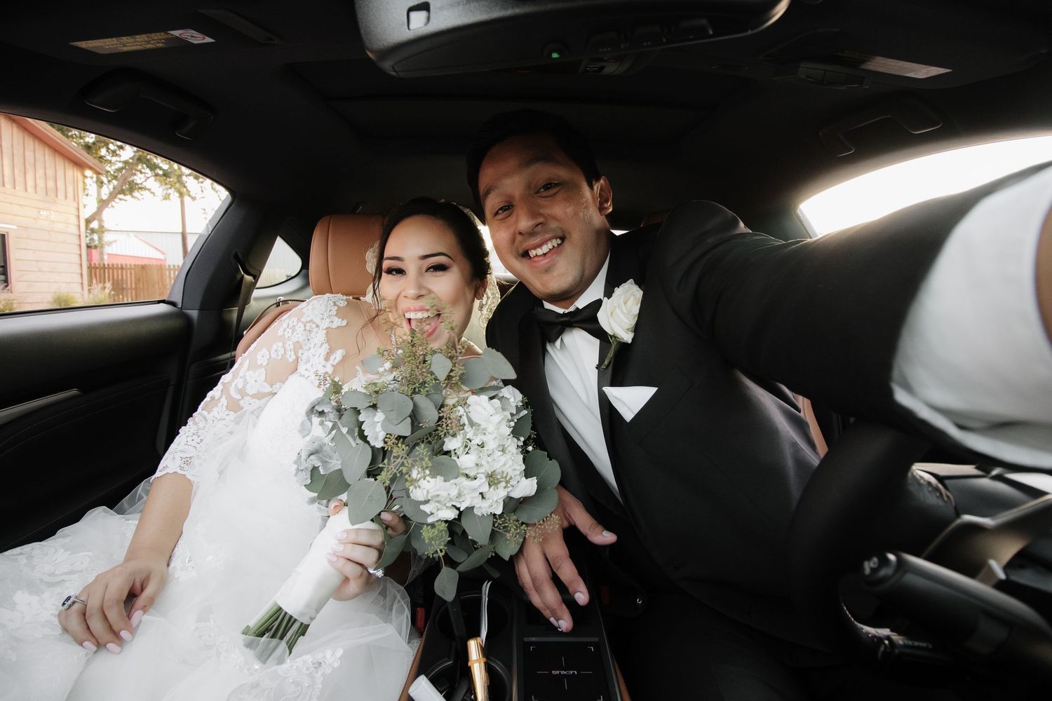 Bride and groom smiling together inside car, bride holding white eucalyptus bouquet.