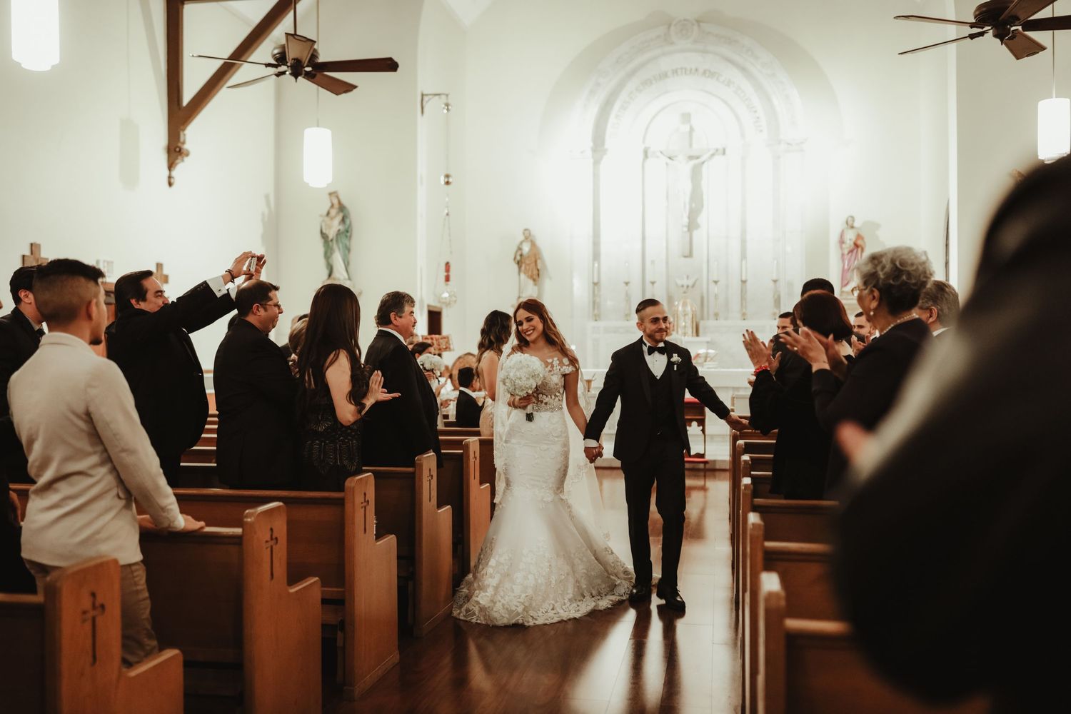 Bride and groom walk joyfully down the aisle of a traditional church surrounded by celebrating guests.