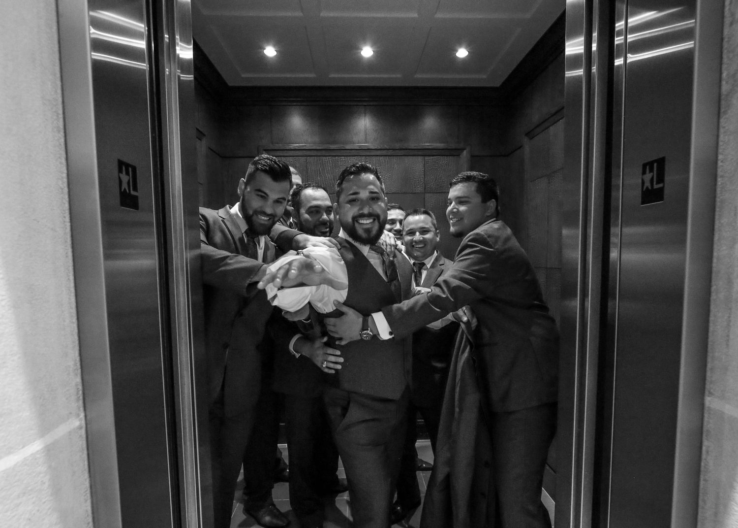 Black and white photo of groom surrounded by groomsmen celebrating in an elevator.
