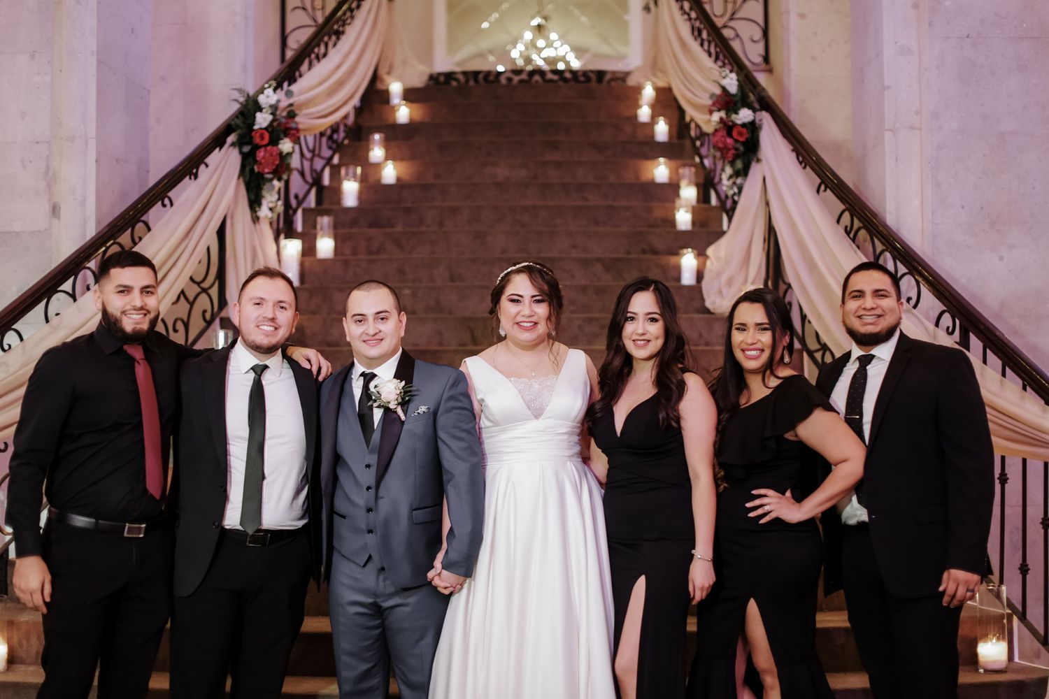 Bride and groom pose with wedding party on grand staircase with floral decorations.