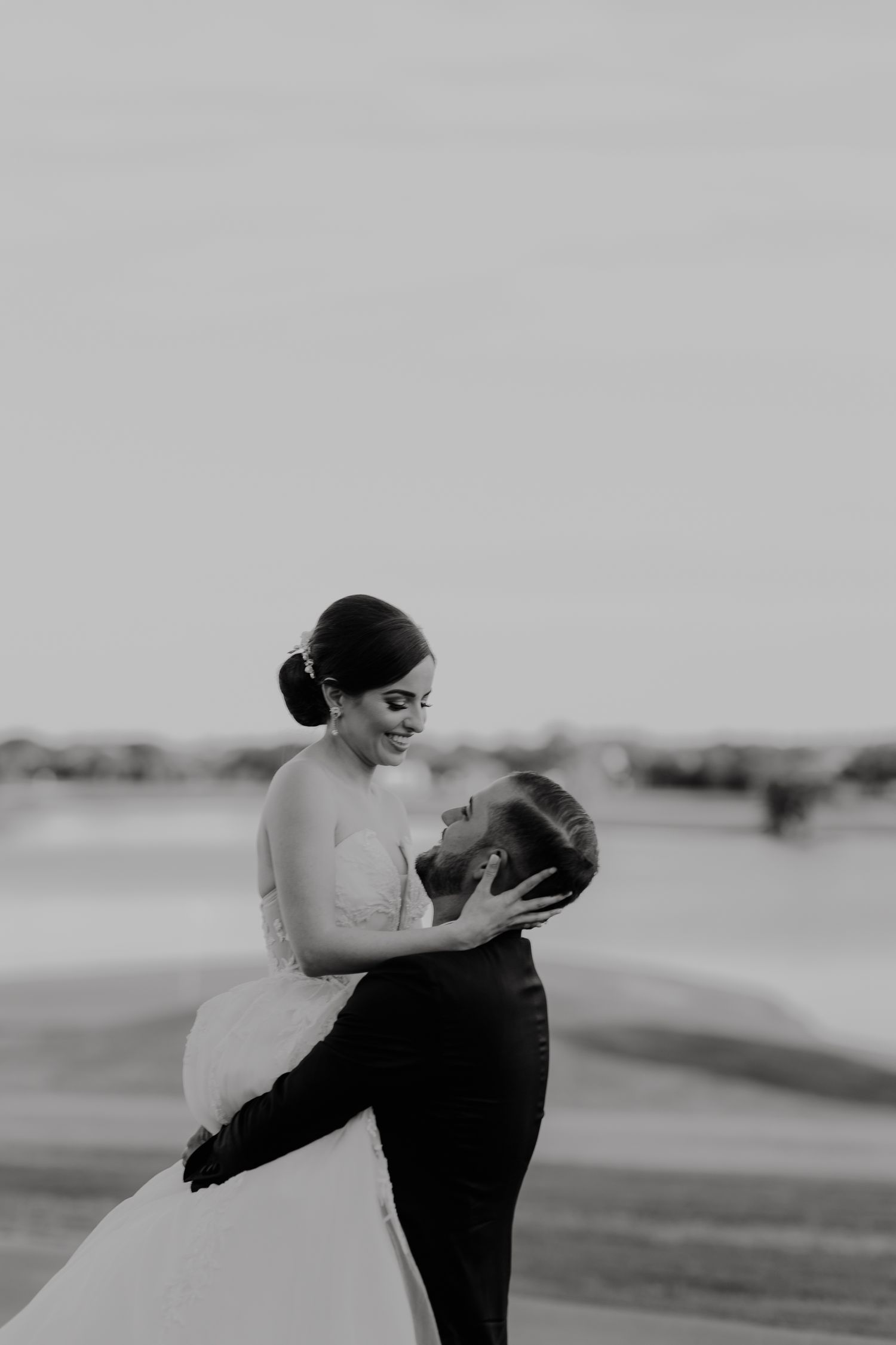 Black and white photo of a groom lifting his bride in a wedding dress outdoors, both smiling joyfully.