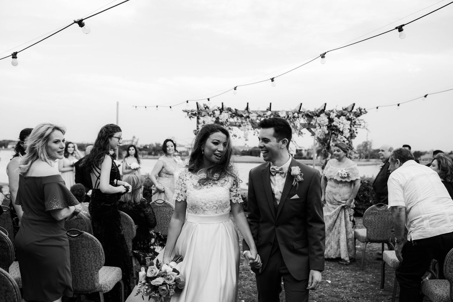Newlyweds walk down the aisle hand in hand at outdoor wedding ceremony with string lights overhead.