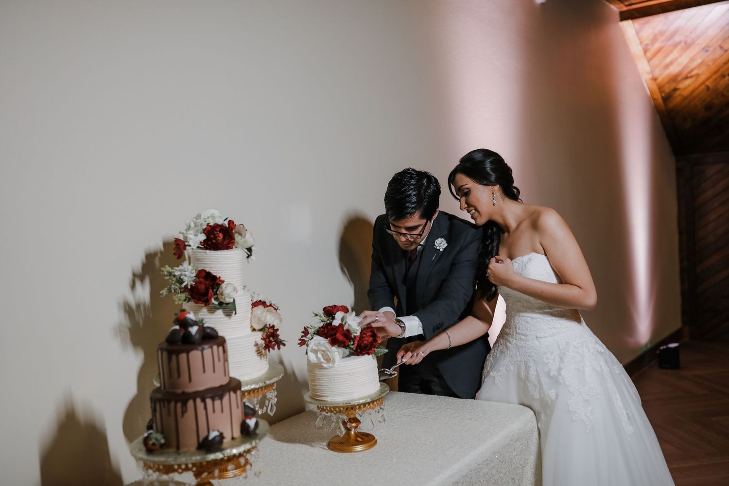 Bride and groom cut elegant wedding cake with red floral decorations at romantic indoor reception.
