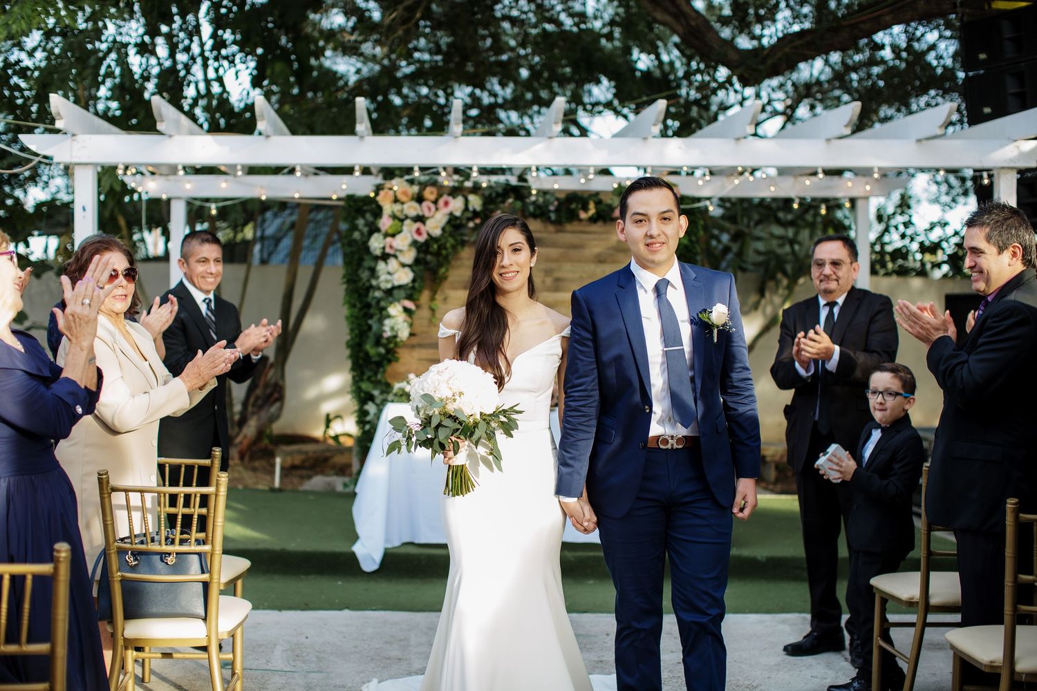 Newlyweds walk down aisle as guests applaud at elegant outdoor garden wedding ceremony.