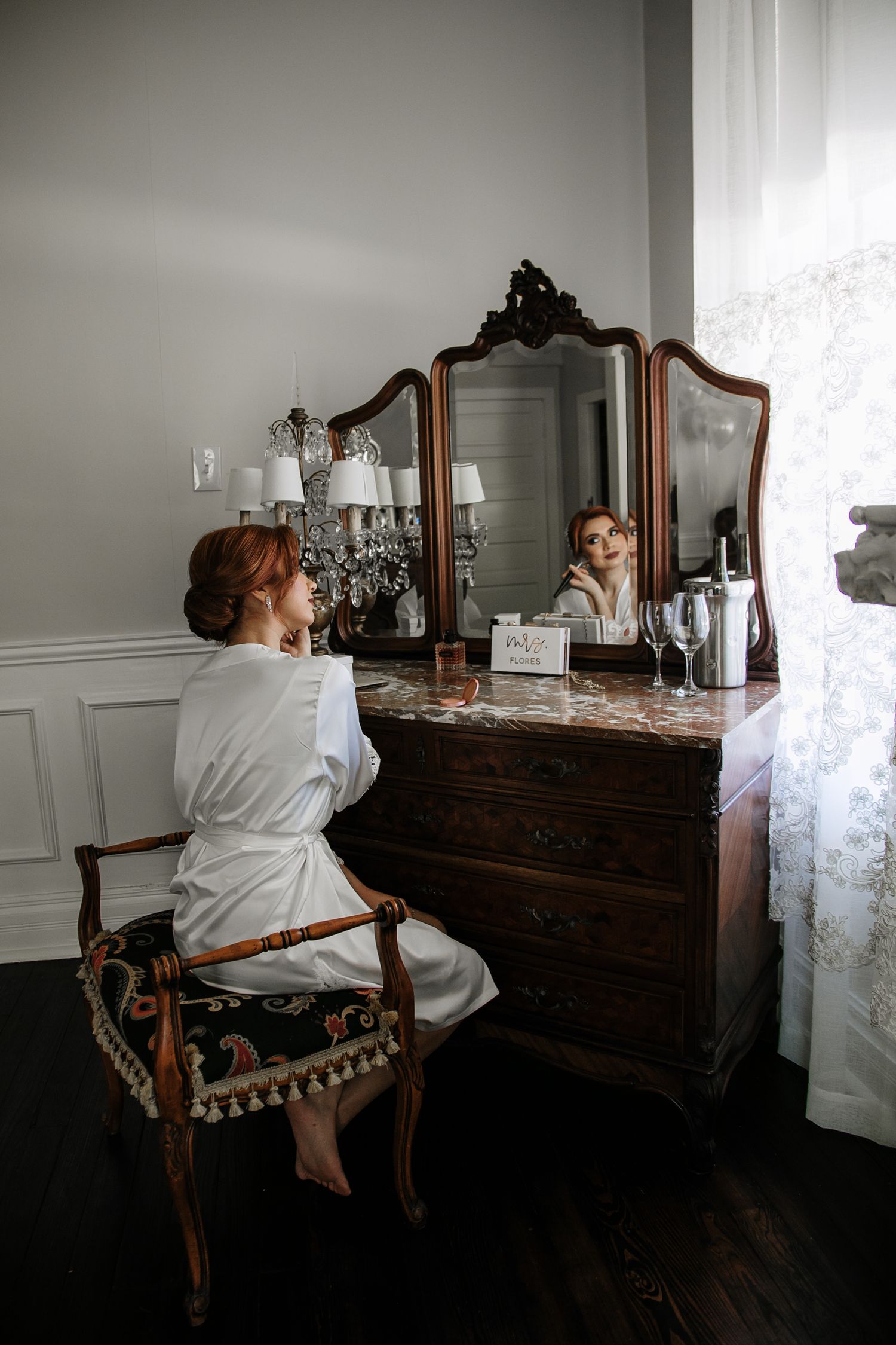 Bride in white robe sits at ornate vintage vanity mirror getting ready on wedding day.