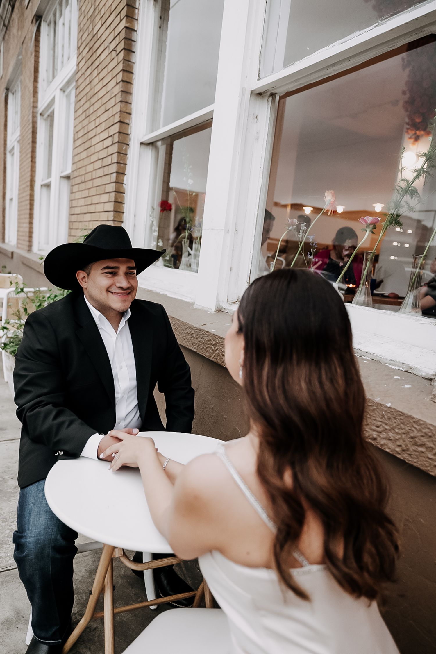 A smiling man in a black cowboy hat and blazer holds hands with a woman at an outdoor cafe table.