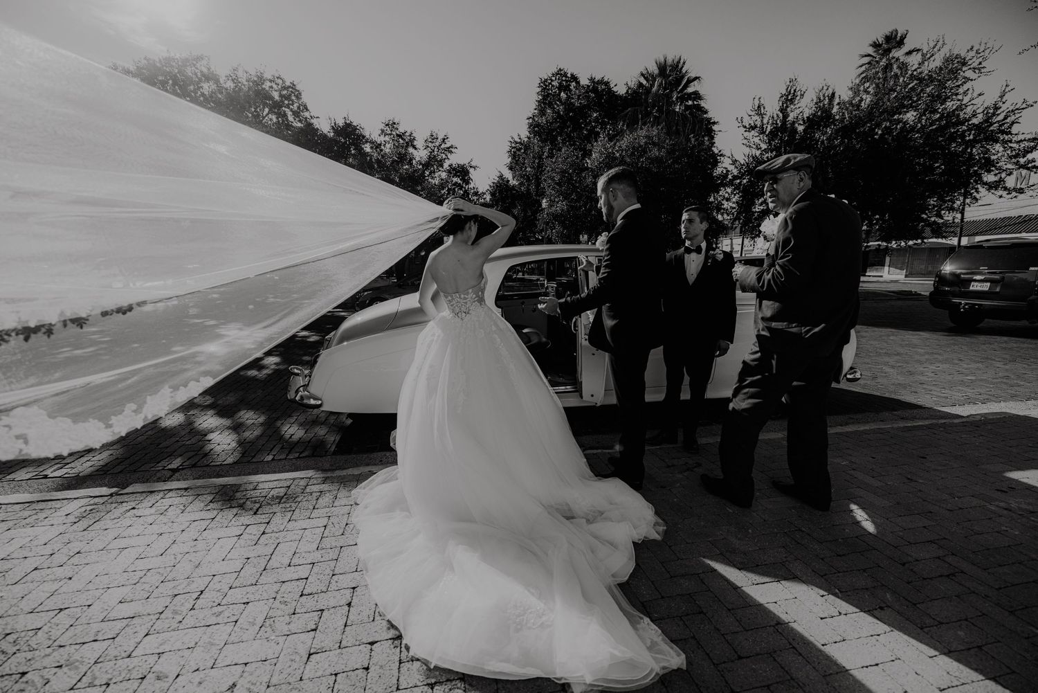 Black and white photo of a bride in a flowing gown and veil arriving at her wedding venue with suited guests nearby.