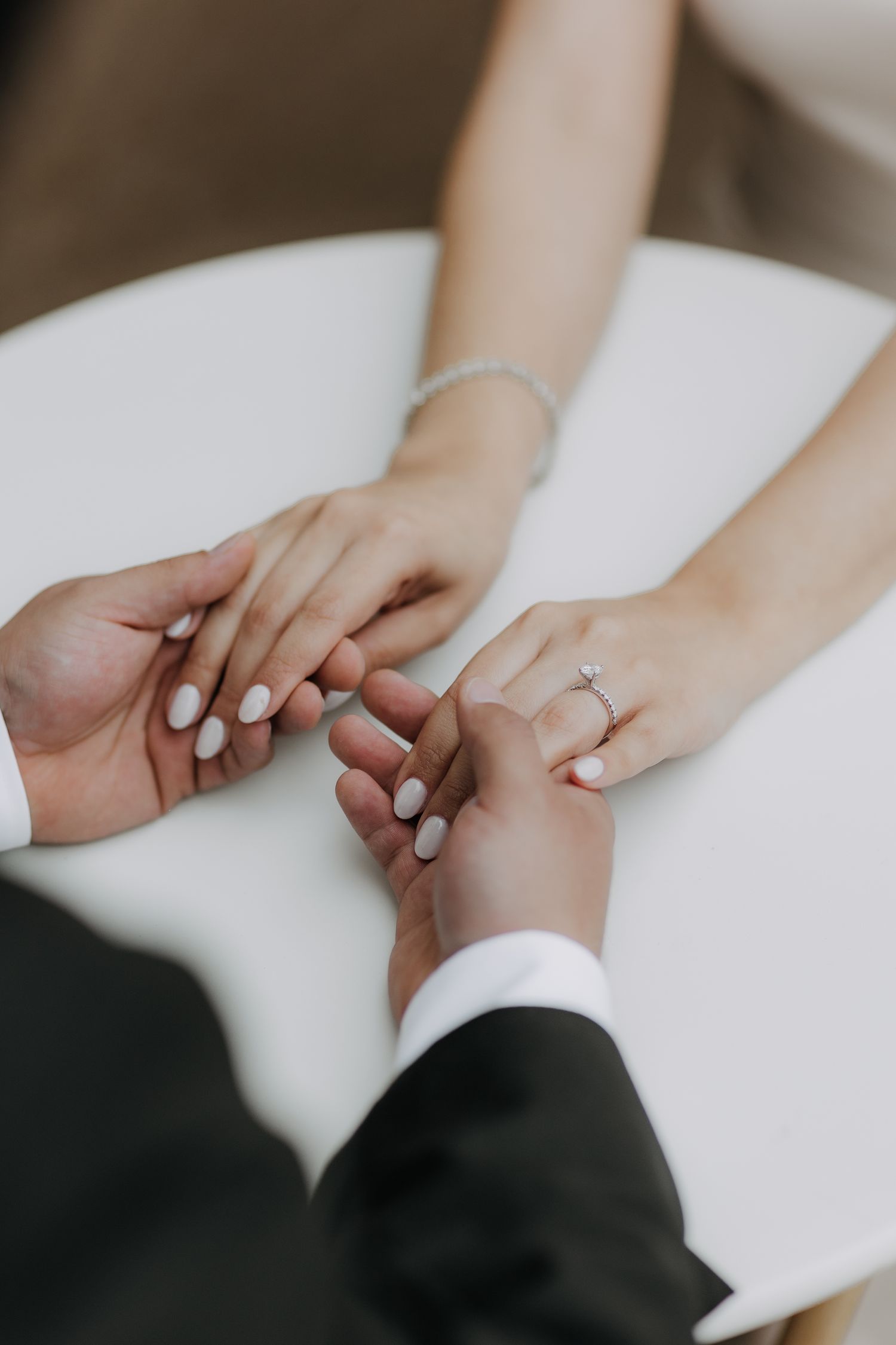 Couple holding hands on white table, bride wearing diamond engagement ring and bracelet, groom in black suit.
