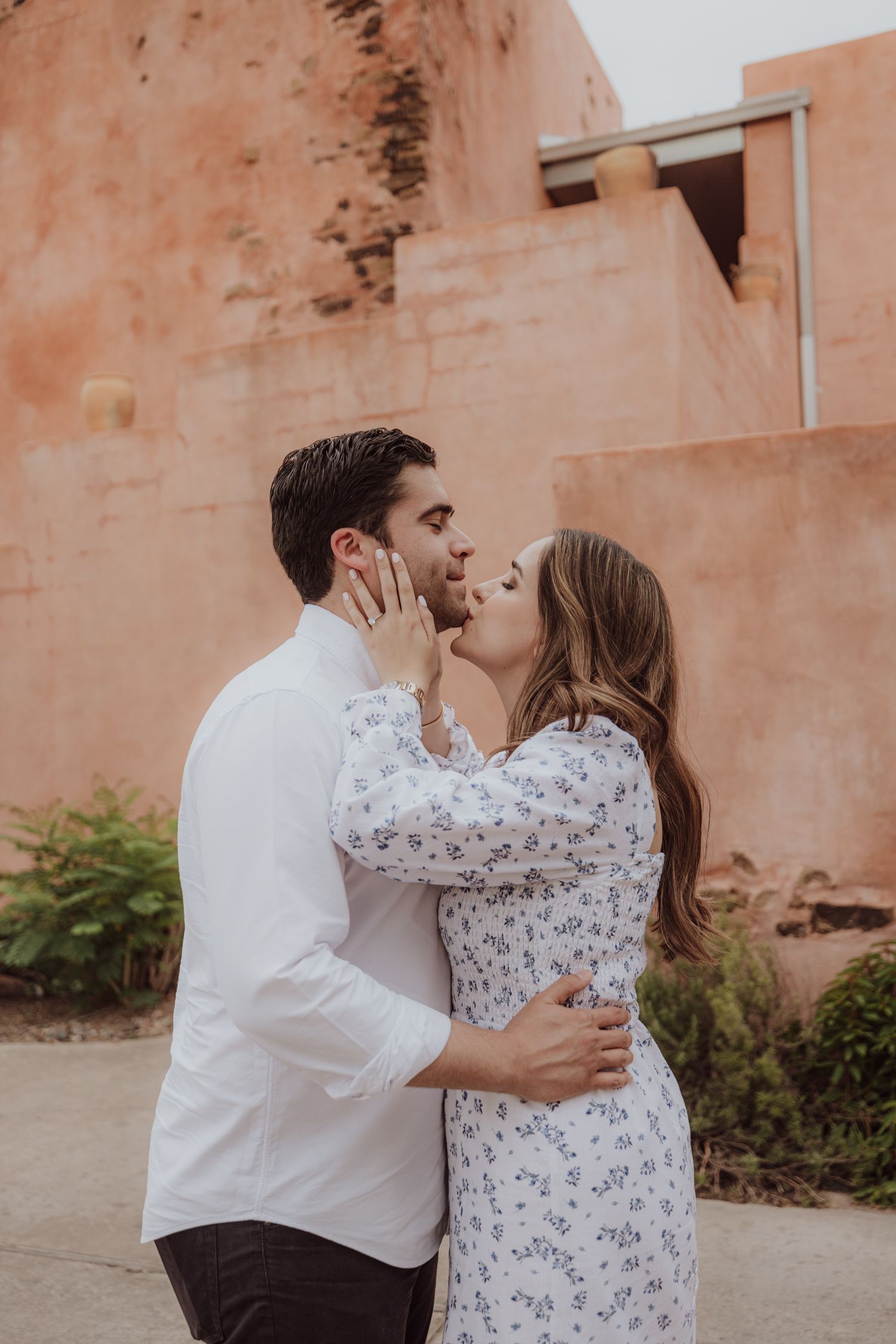 Couple sharing a romantic kiss outdoors near terracotta adobe walls, woman in floral dress holding mans face.