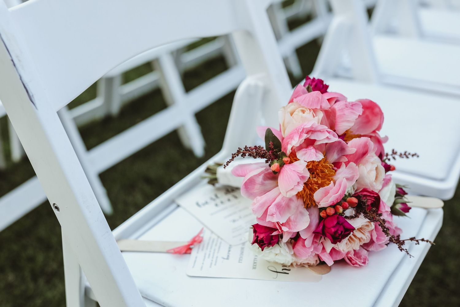 Vibrant pink and red bridal bouquet with peonies rests on a white folding chair at an outdoor ceremony.