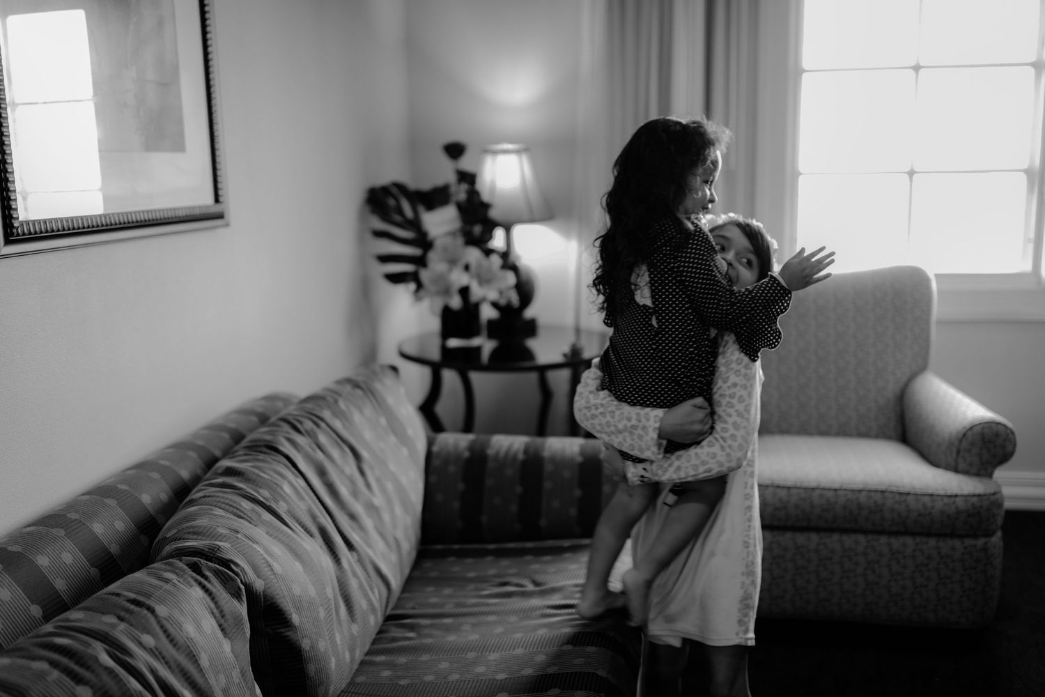 Black and white photo of bride embracing child by window in hotel room.