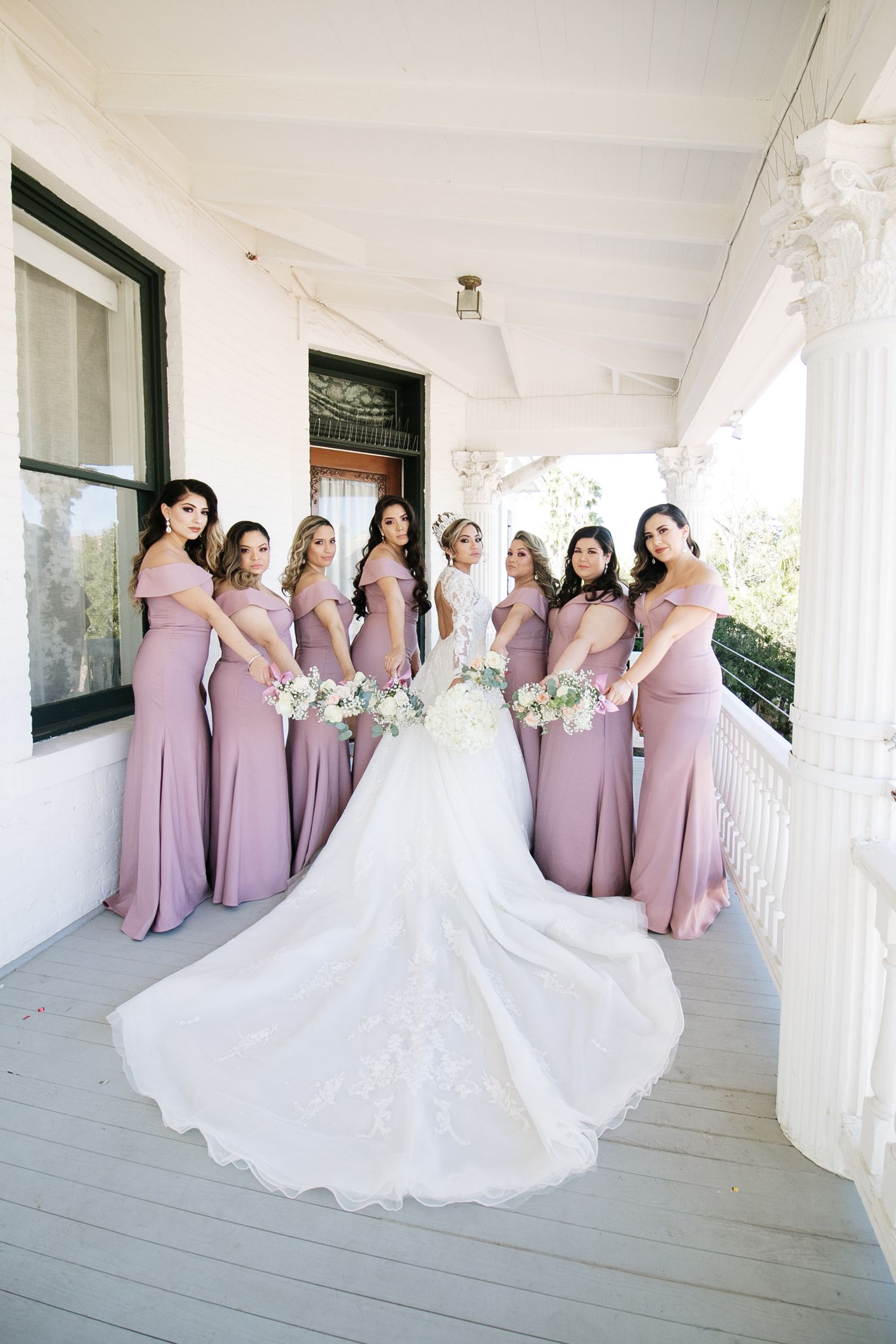 Bride with long lace train poses with bridesmaids in mauve off-shoulder gowns on white colonial porch.