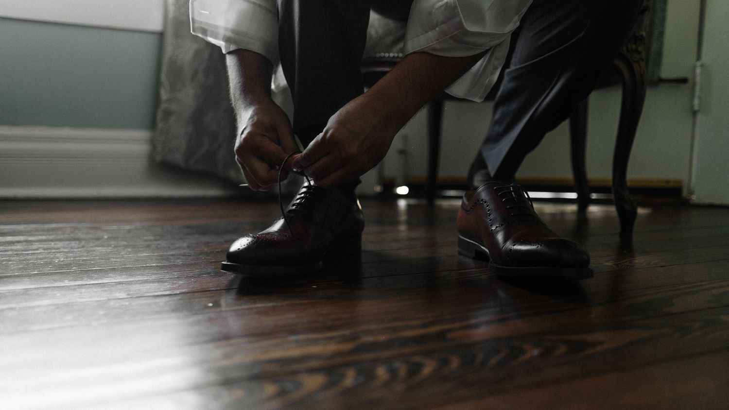 Man tying dark brown Oxford dress shoes on hardwood floor in formal attire.