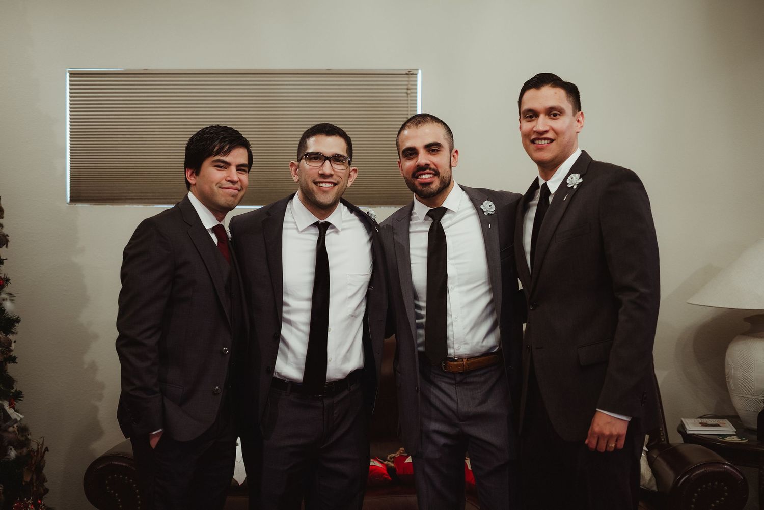 Four groomsmen in dark suits and ties smiling together indoors at wedding.