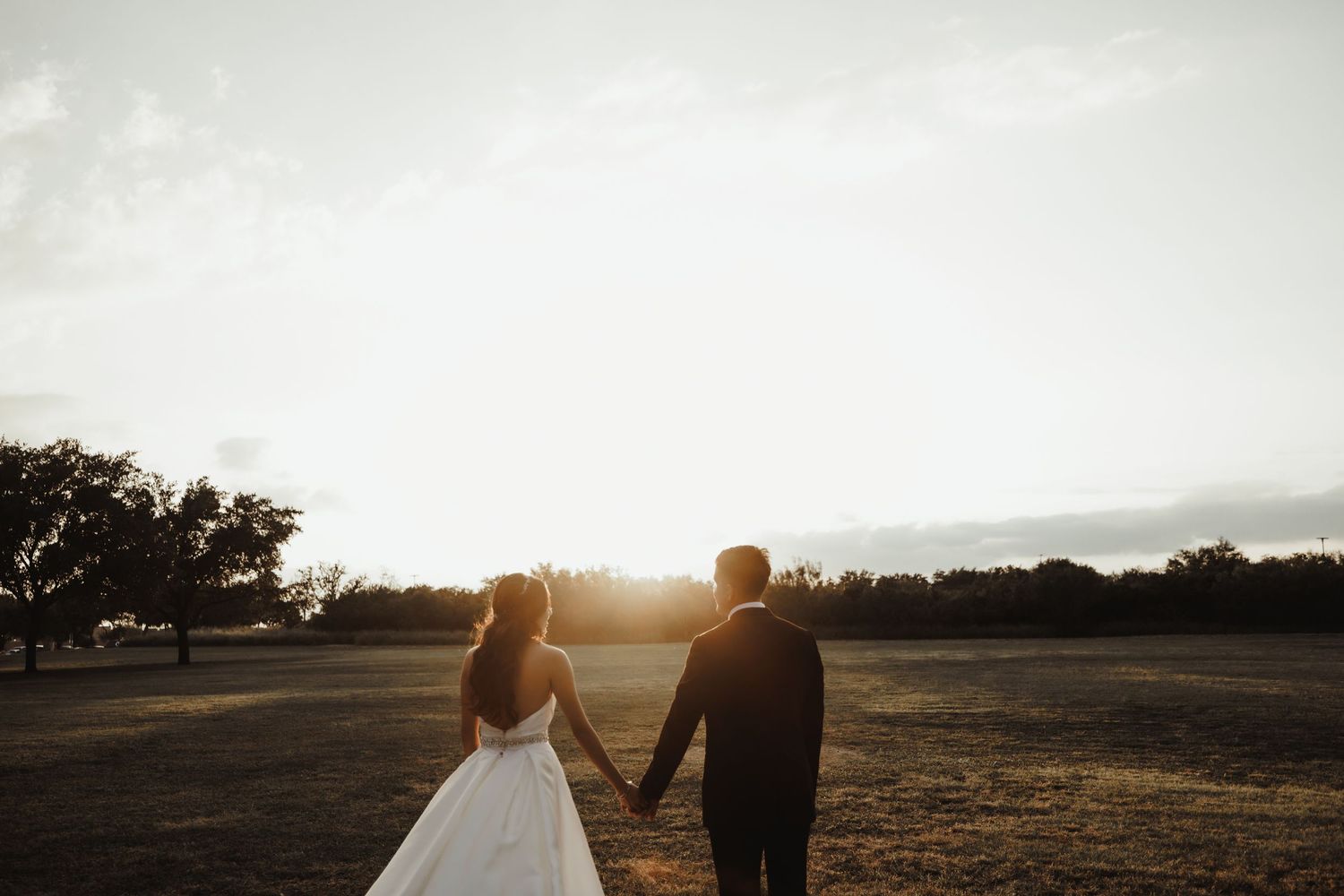 A bride and groom hold hands walking toward a golden sunset across an open field on their wedding day.