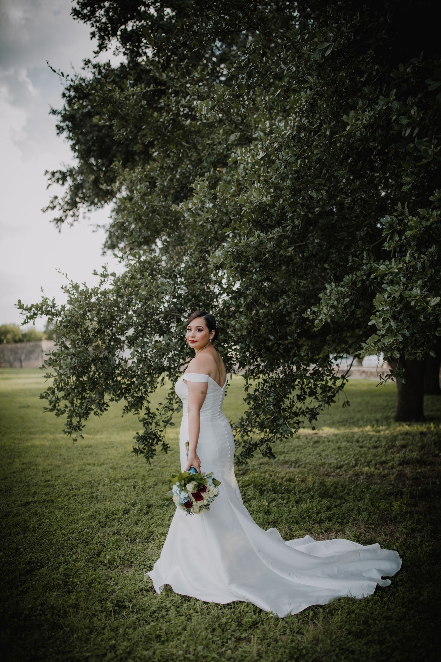Elegant bride in a fitted off-shoulder white gown with a long train standing beneath a large oak tree.