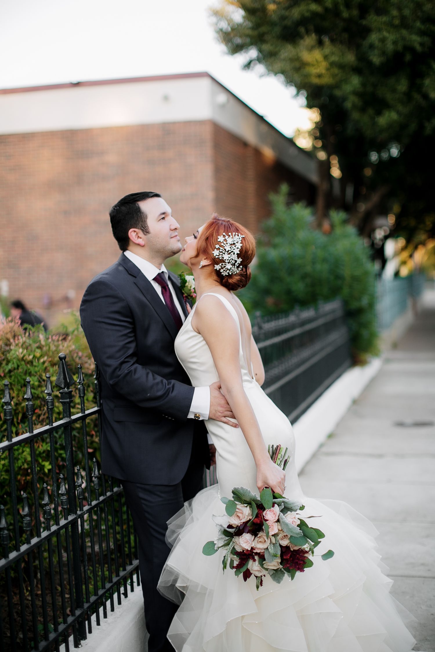 Groom kissing bride on cheek outdoors, bride holding bouquet of red and pink roses on city sidewalk.