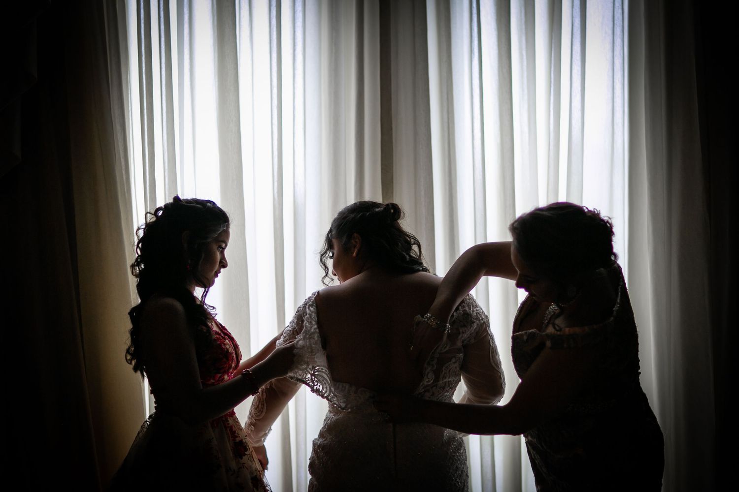Silhouette of bride being helped into wedding dress by two women against curtained window.