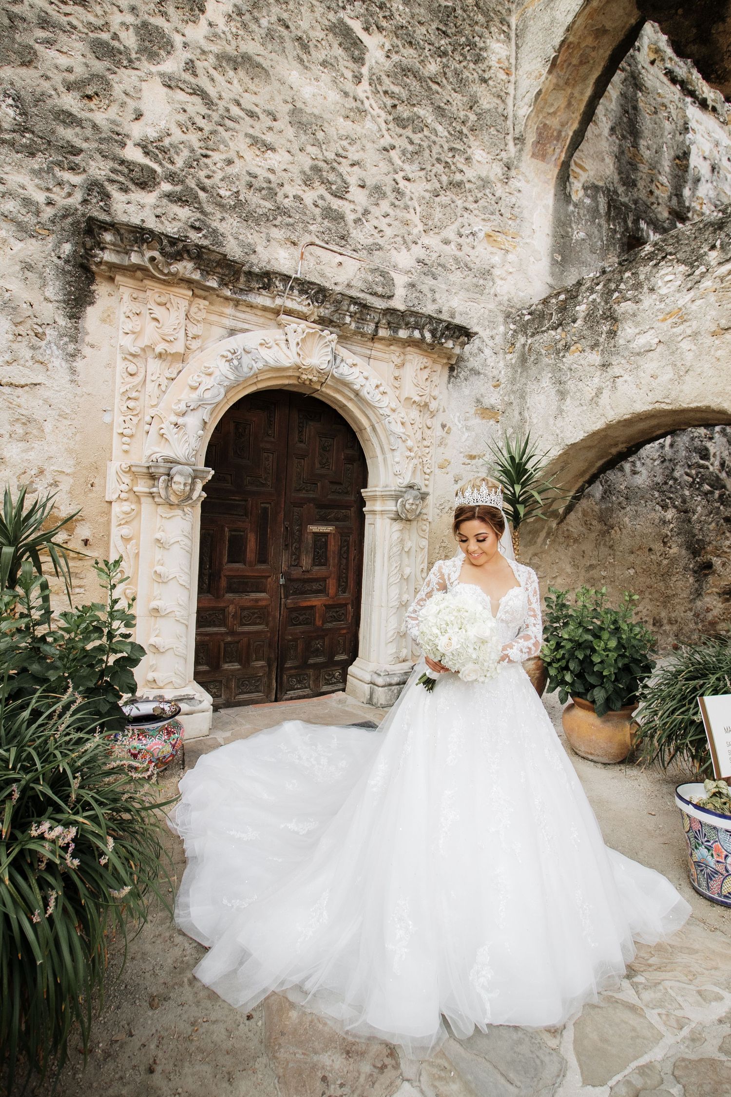 Bride in ballgown with long train poses before ornate wooden door of historic stone church.