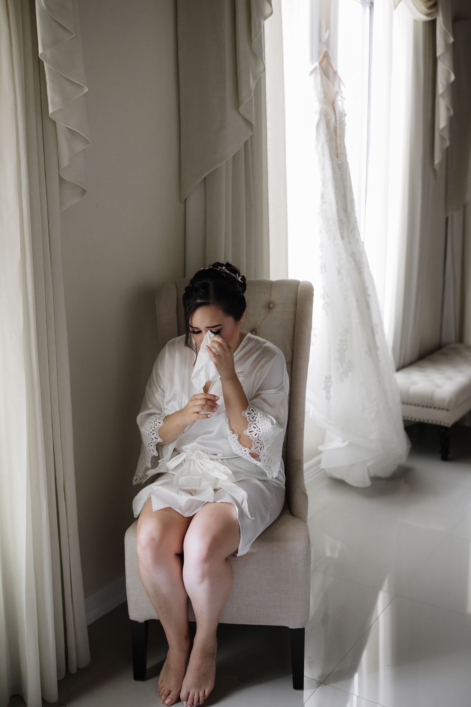Emotional bride in white robe dabs tears while seated in armchair next to her wedding dress by window.
