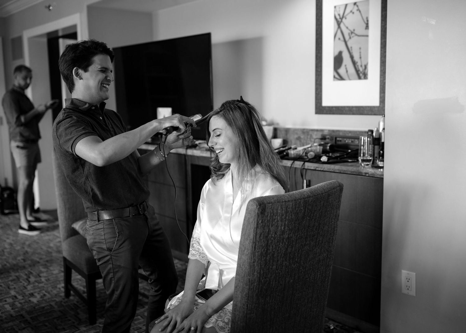 Black and white photo of hairstylist styling smiling bride's hair in a hotel room.