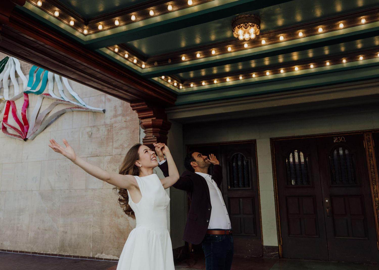 Bride in white dress and groom dancing joyfully outside a vintage theater with marquee lights.